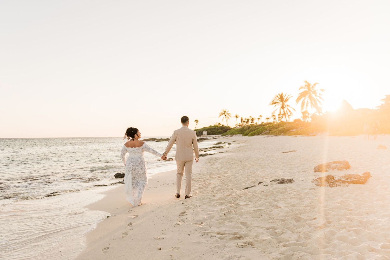 Xcaret Arte Wedding - Couple in wedding attire holding hands walking along a tropical beach at golden sunset with palm trees.