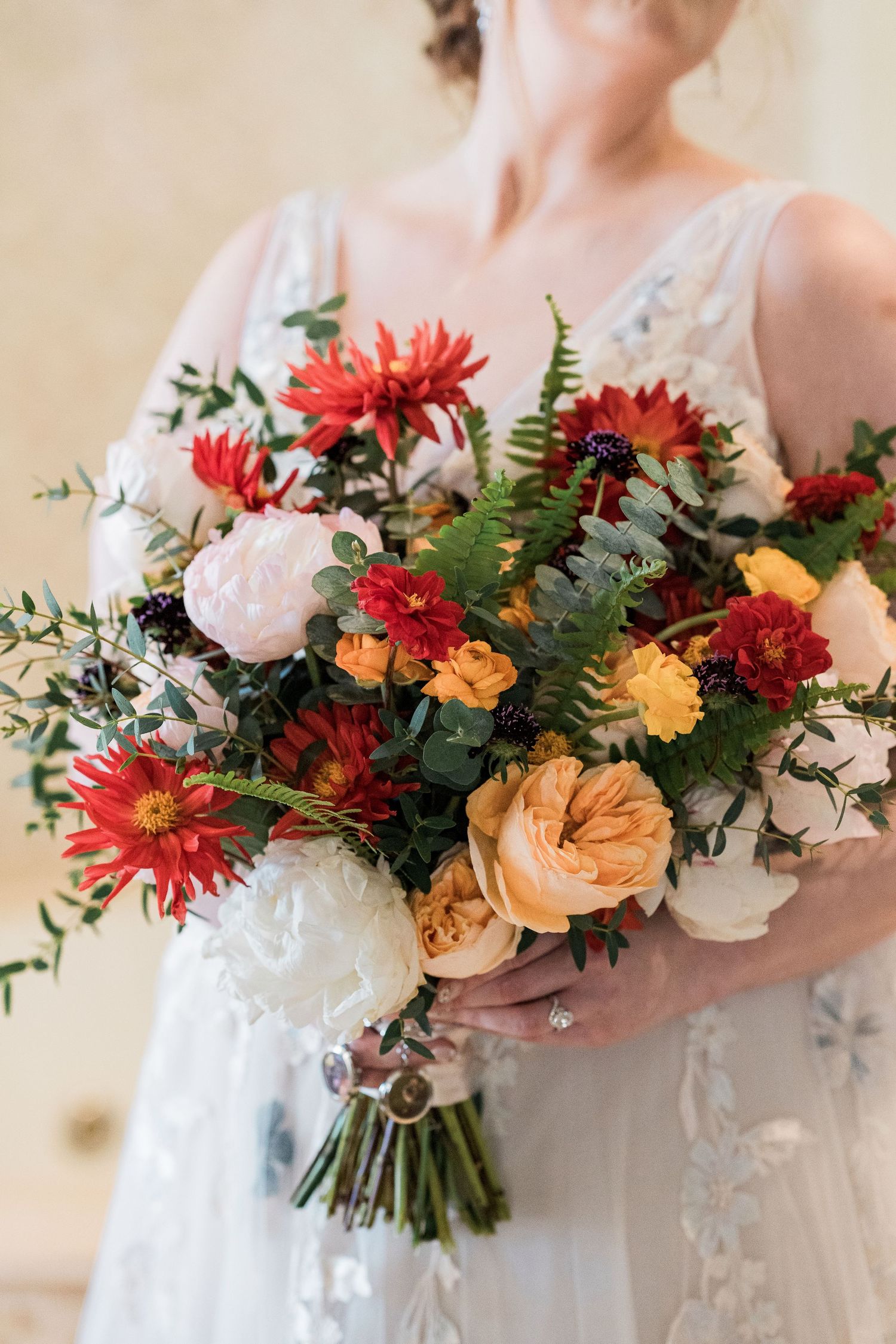 Bride in white lace gown holding a lush wedding bouquet with red, orange, and white blooms and greenery.