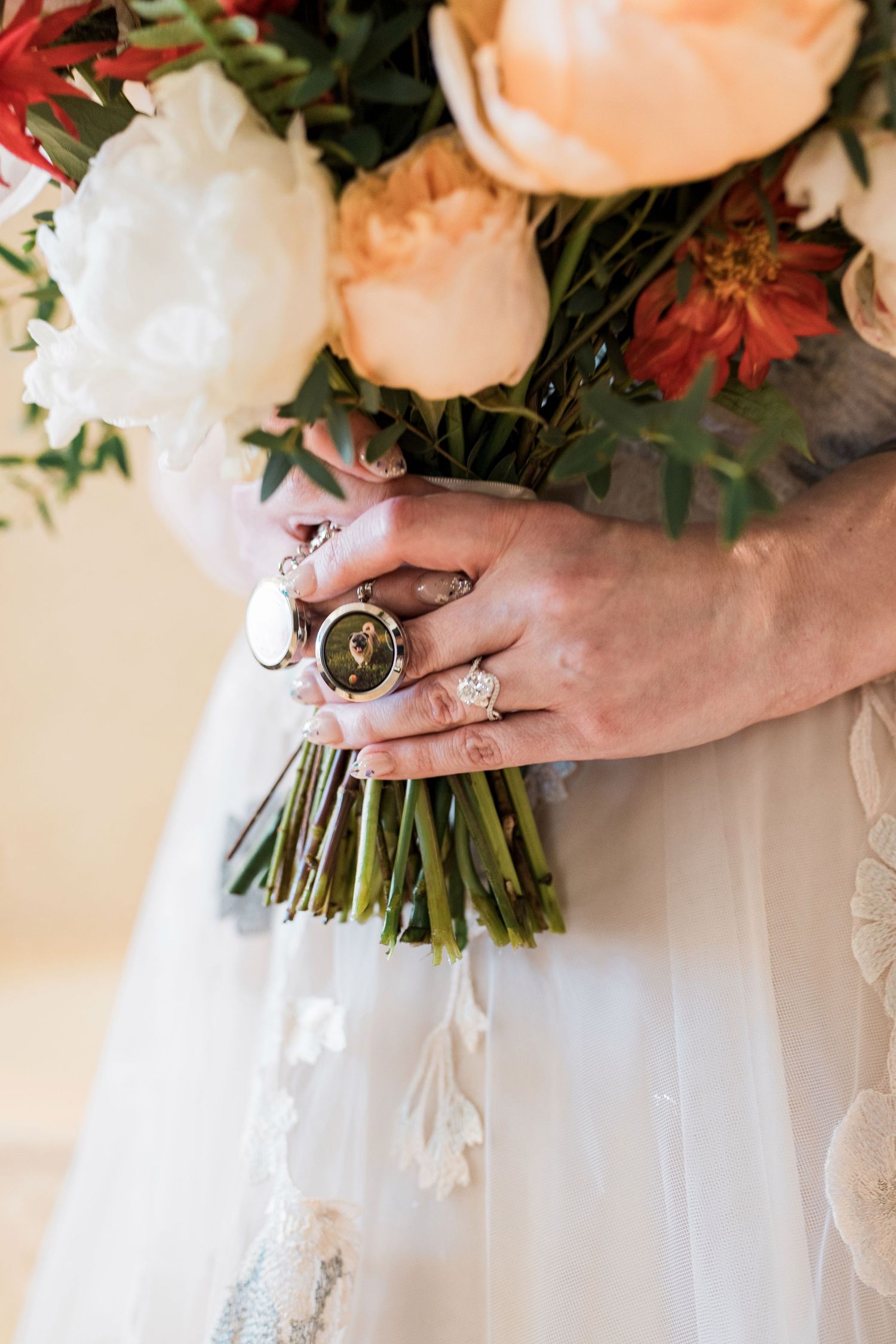 Bride holding a bouquet of peach, white, and red flowers, wearing a diamond engagement ring and a locket charm.