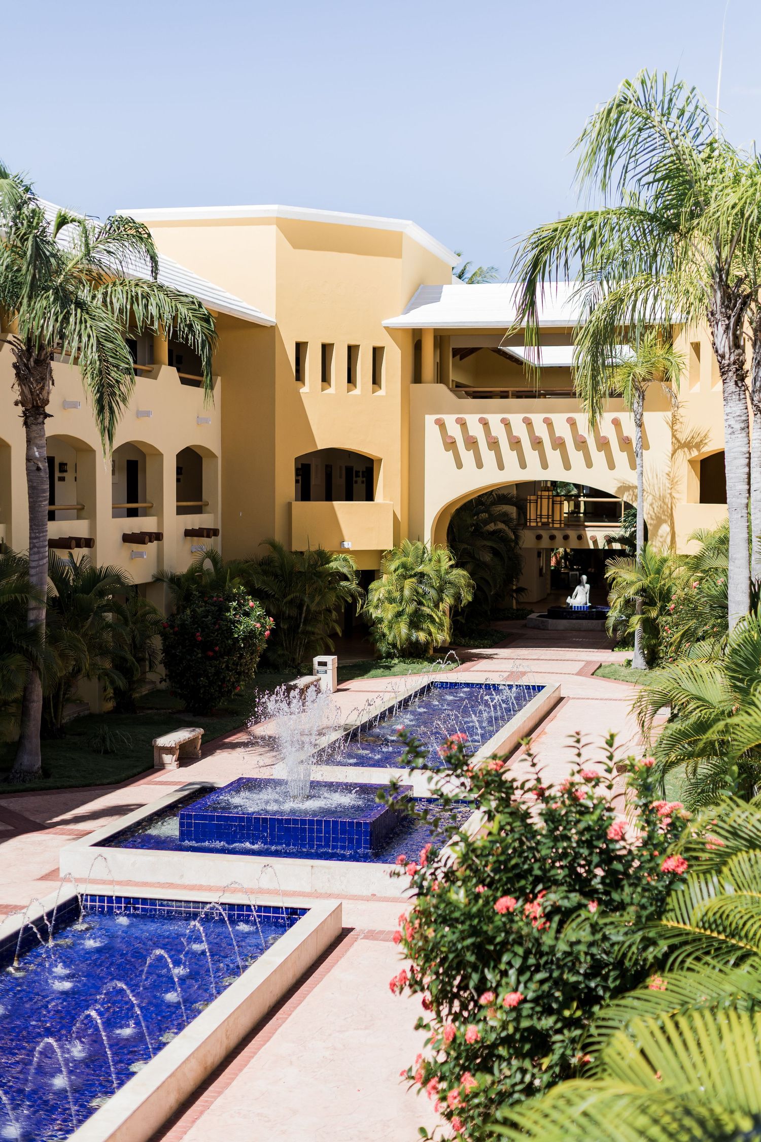 Barcelo Maya Grand Resort -Tropical resort courtyard with blue tile fountains, palm trees, and yellow Mediterranean-style architecture.