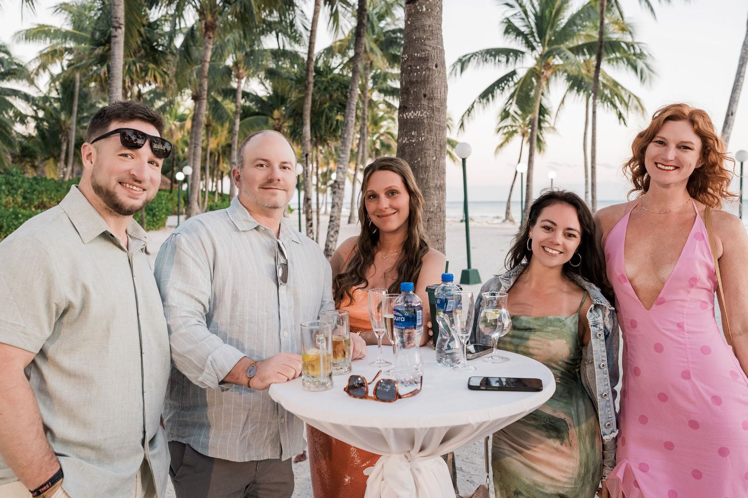 Five friends smiling at a tropical beach party with palm trees, cocktails, and warm sunset lighting.