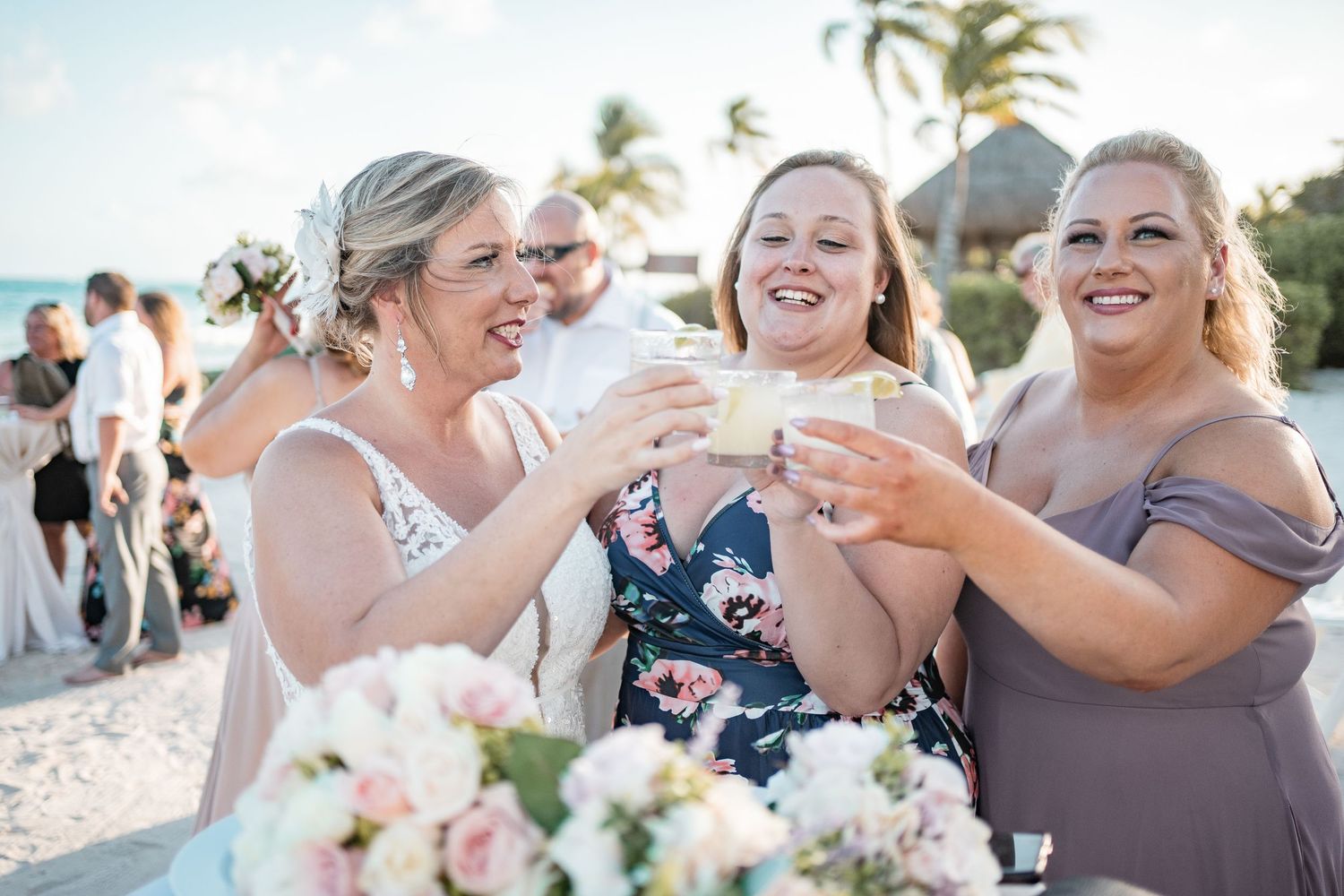 Bride and bridesmaids joyfully toast with drinks at outdoor beach wedding reception celebration.