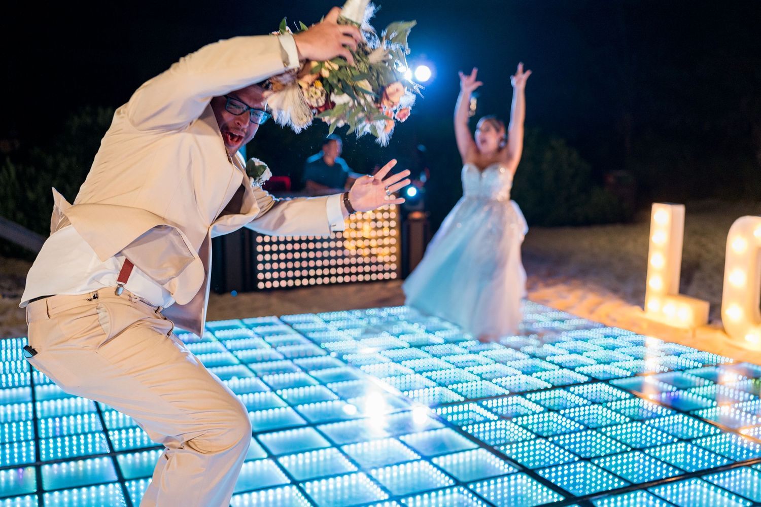Groom tossing bouquet on illuminated dance floor as bride poses at outdoor beach wedding reception.
