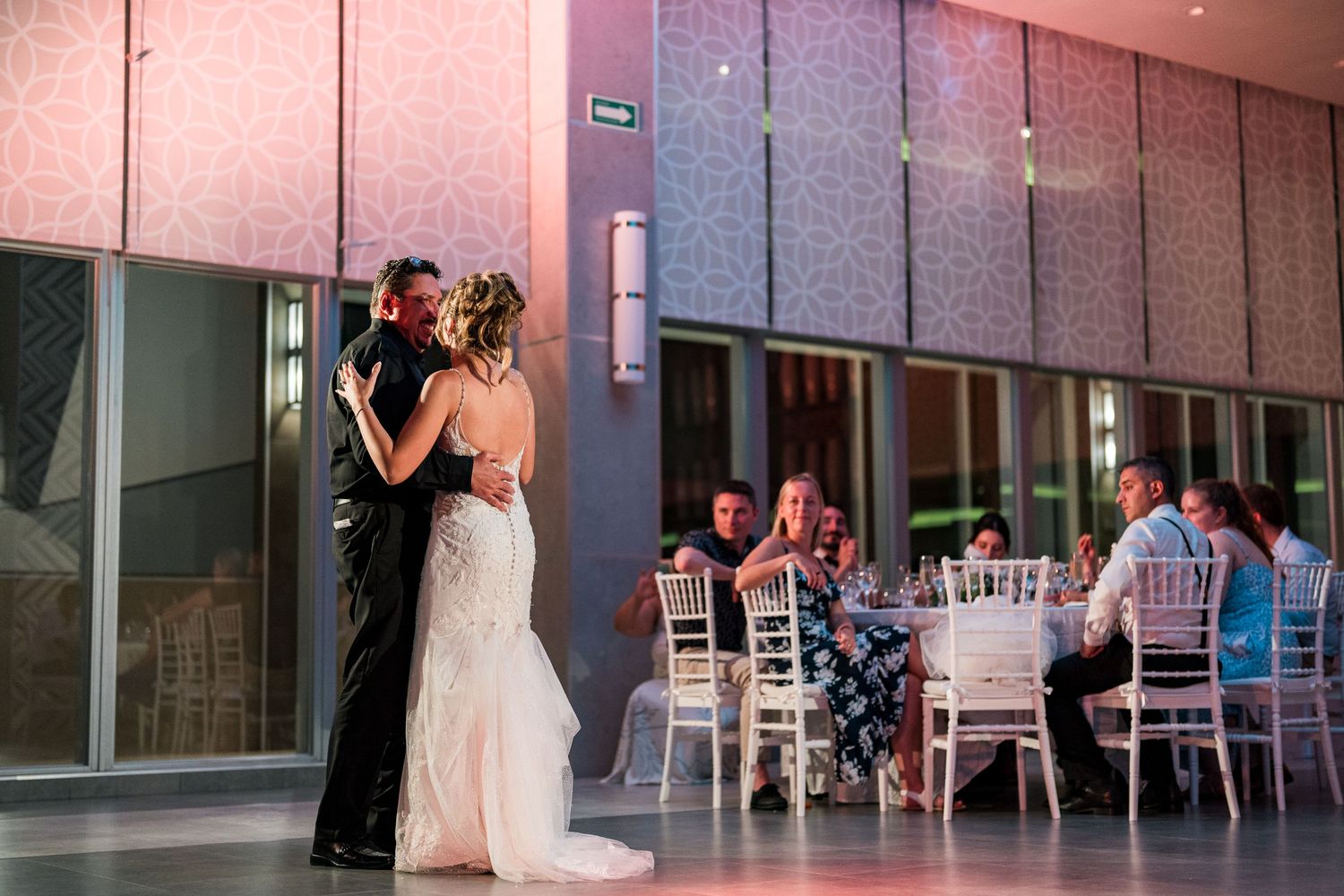 Bride and groom share their first dance at elegant wedding reception with guests seated at white chairs.