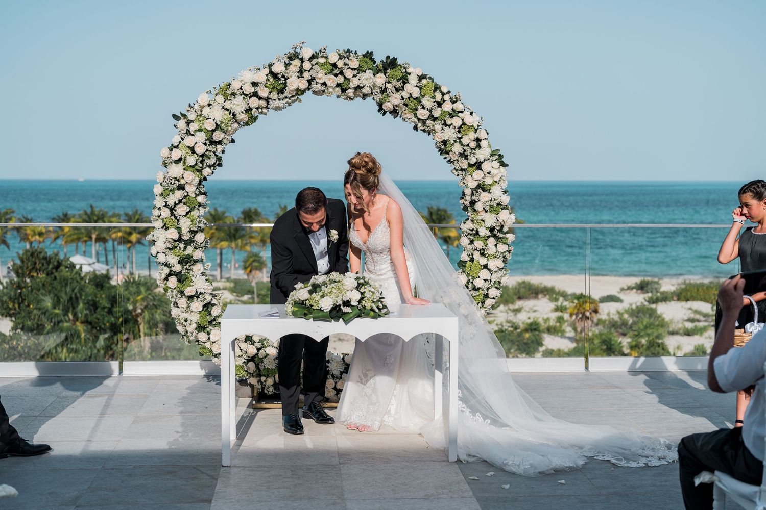 Bride and groom sign marriage certificate under circular floral arch at oceanfront venue.