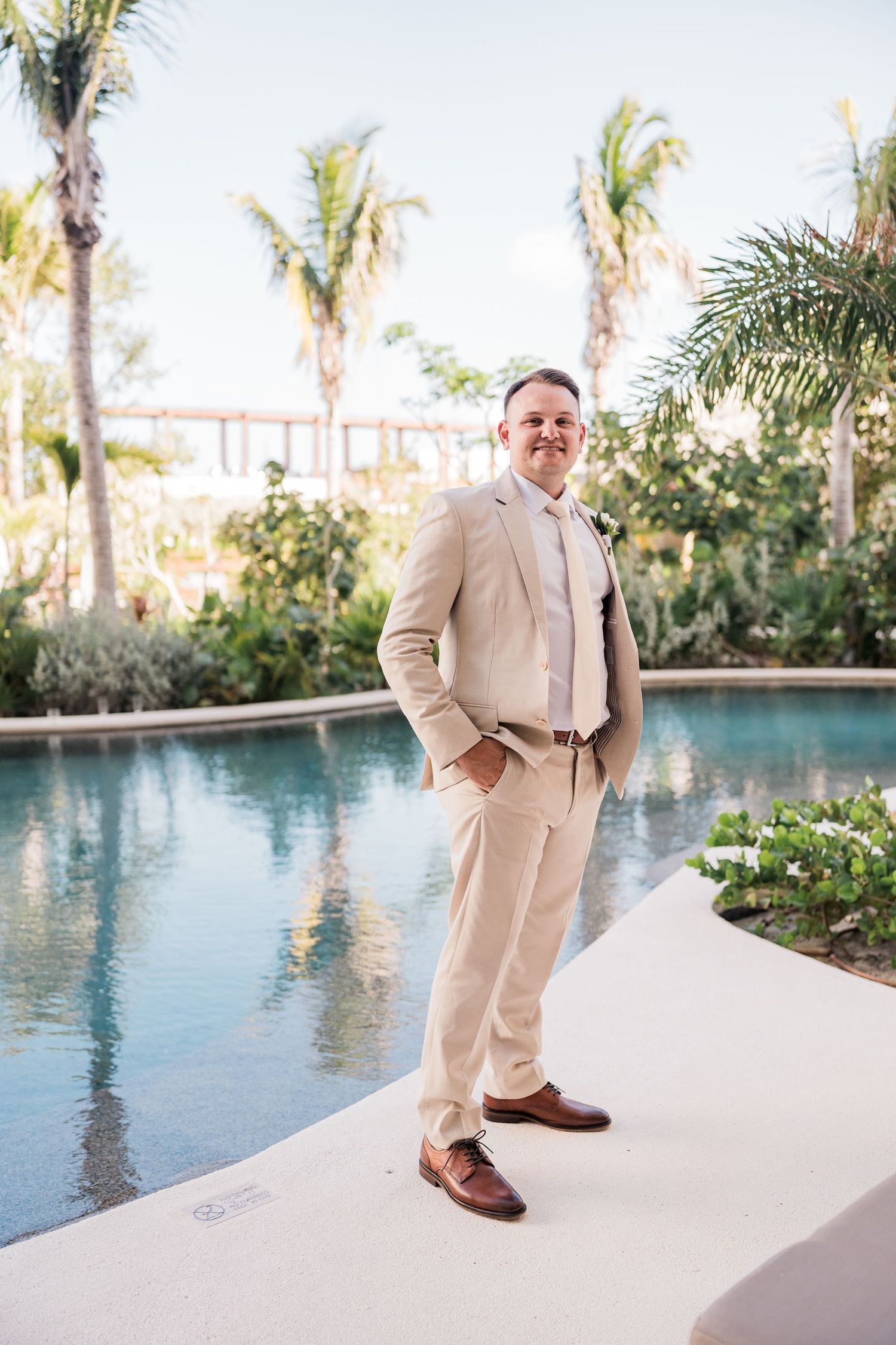 Groom in beige suit standing by resort pool with palm trees at tropical destination wedding.