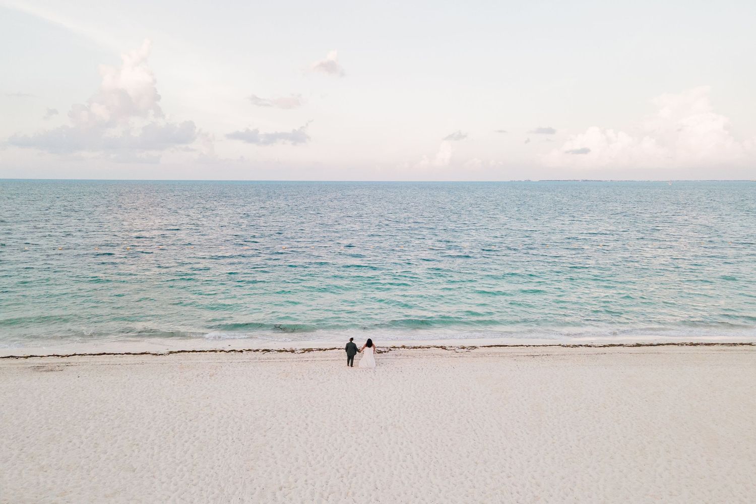 Aerial view of two tiny figures in wedding attire standing at the edge of a vast white tropical beach.