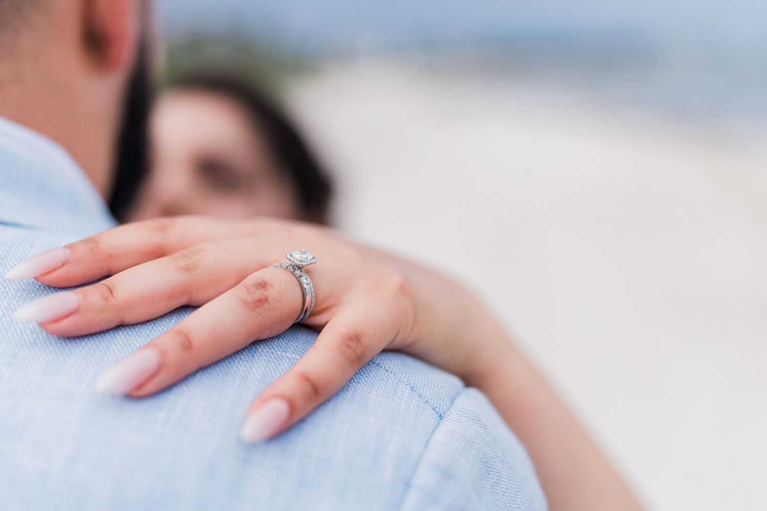 Close-up of a diamond engagement ring on bride's hand resting on groom's light blue suit jacket at beach wedding.