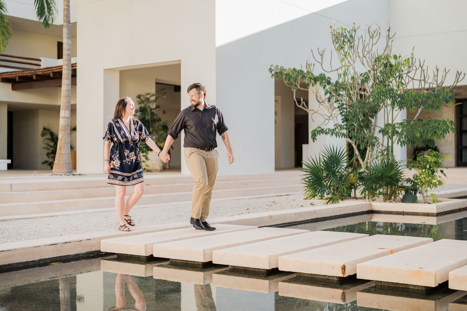Secrets Akumal Wedding - Couple holding hands and walking on stepping stones over a reflective pool at a modern resort.