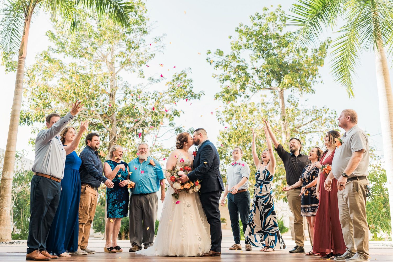 Secrets Akumal Wedding - Bride and groom kiss surrounded by cheering guests tossing confetti at an outdoor wedding ceremony with palm trees.