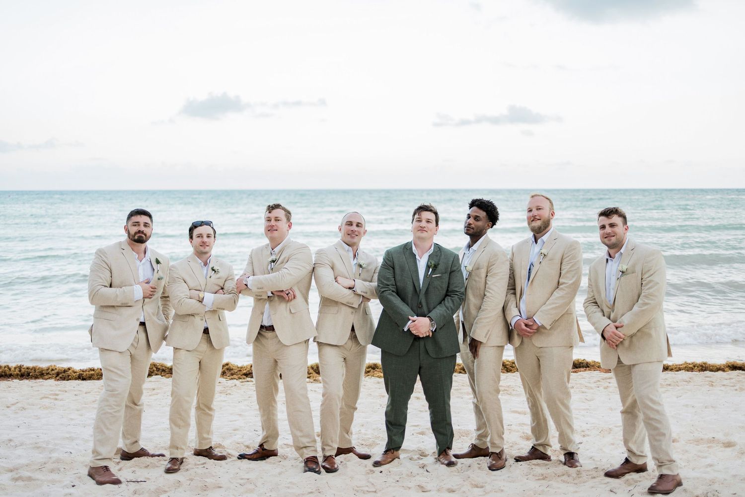 Groom in green suit stands with seven groomsmen in tan suits on a beach with ocean backdrop.