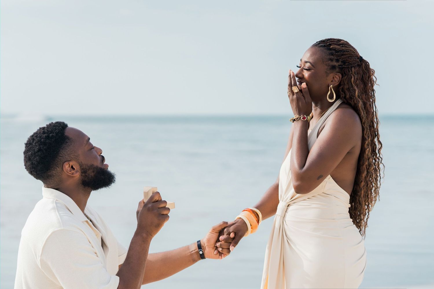 Man proposing to woman on beach, she reacts with joy and surprise, ocean backdrop.