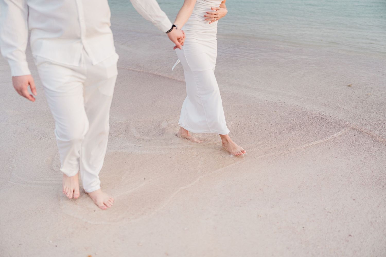 Couple in white outfits holding hands walking barefoot on a sandy beach with calm turquoise water.