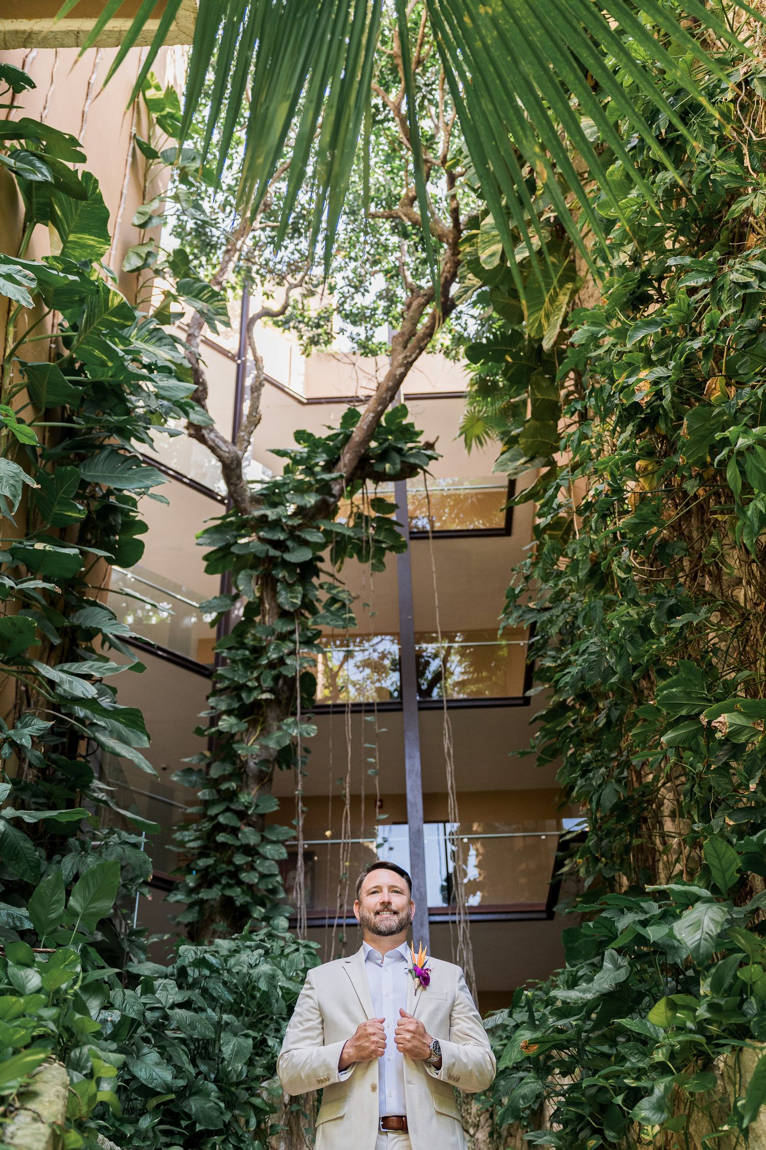 Hotel Xcaret México Wedding  Groom in beige suit stands confidently among lush tropical greenery in an atrium, wearing a floral boutonniere.
