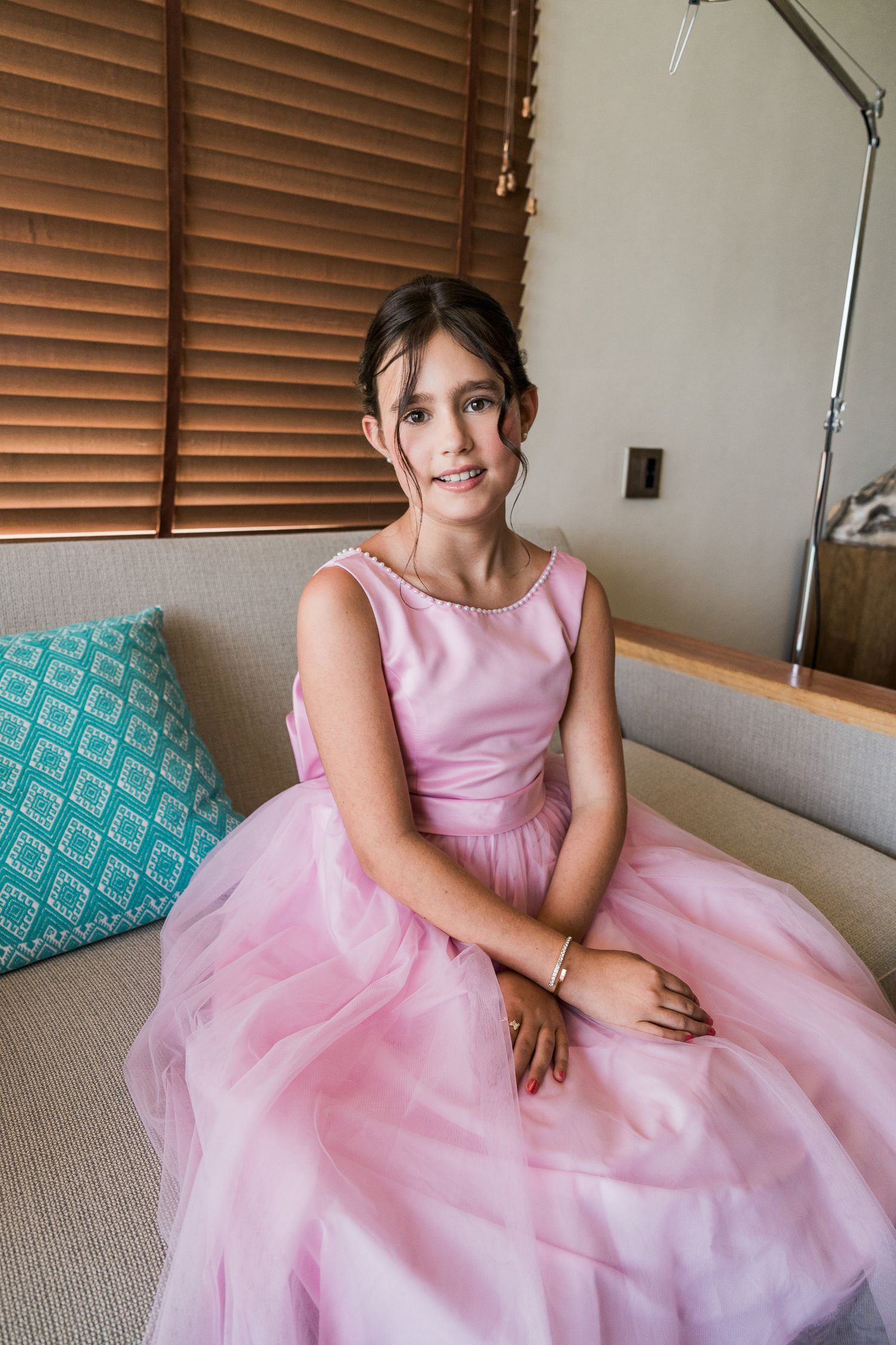 Young girl in pink tulle dress sitting on sofa with teal pillow, smiling at camera near wooden blinds.