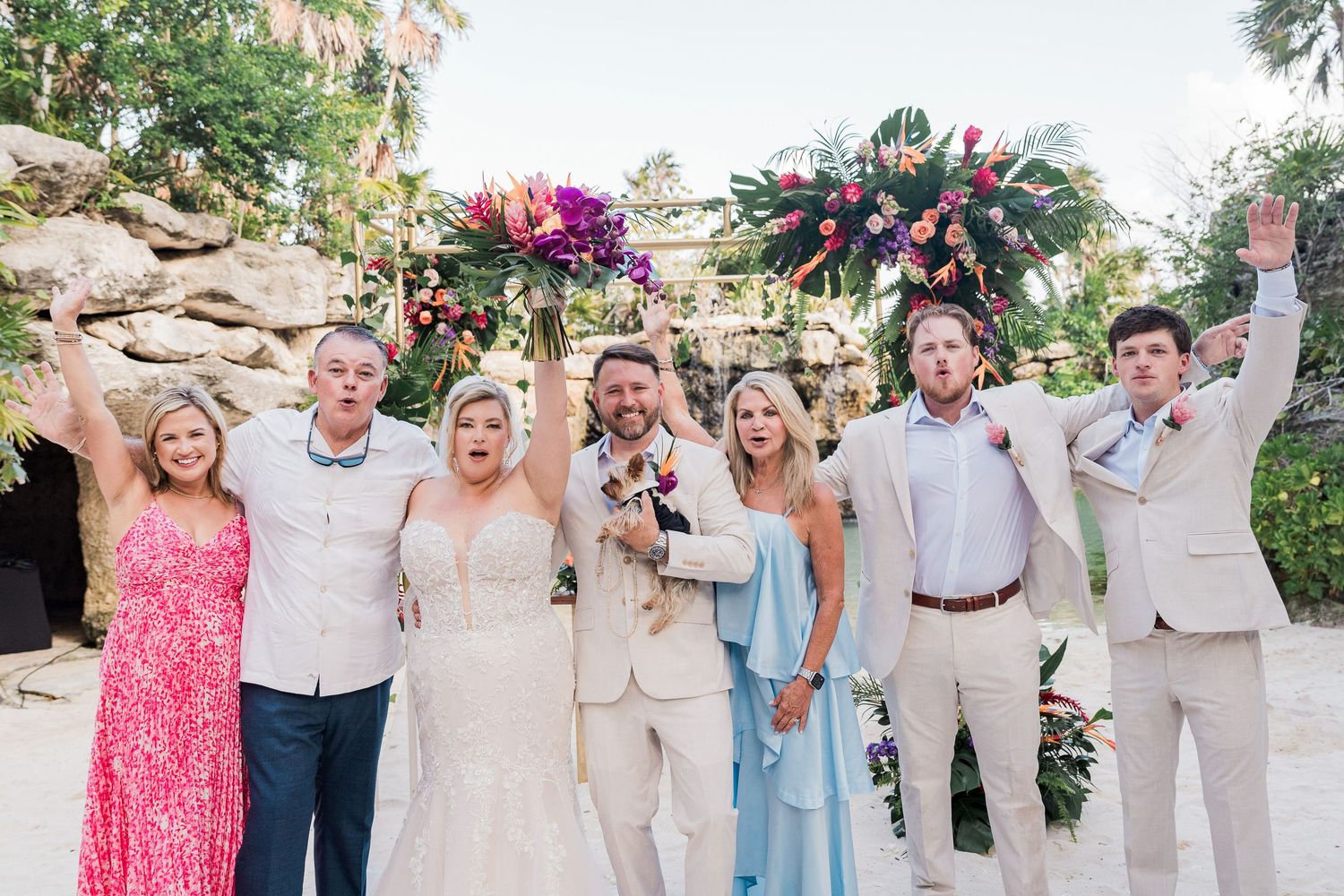 Wedding party celebrating outdoors under a colorful floral arch, guests in formal attire cheering joyfully.
