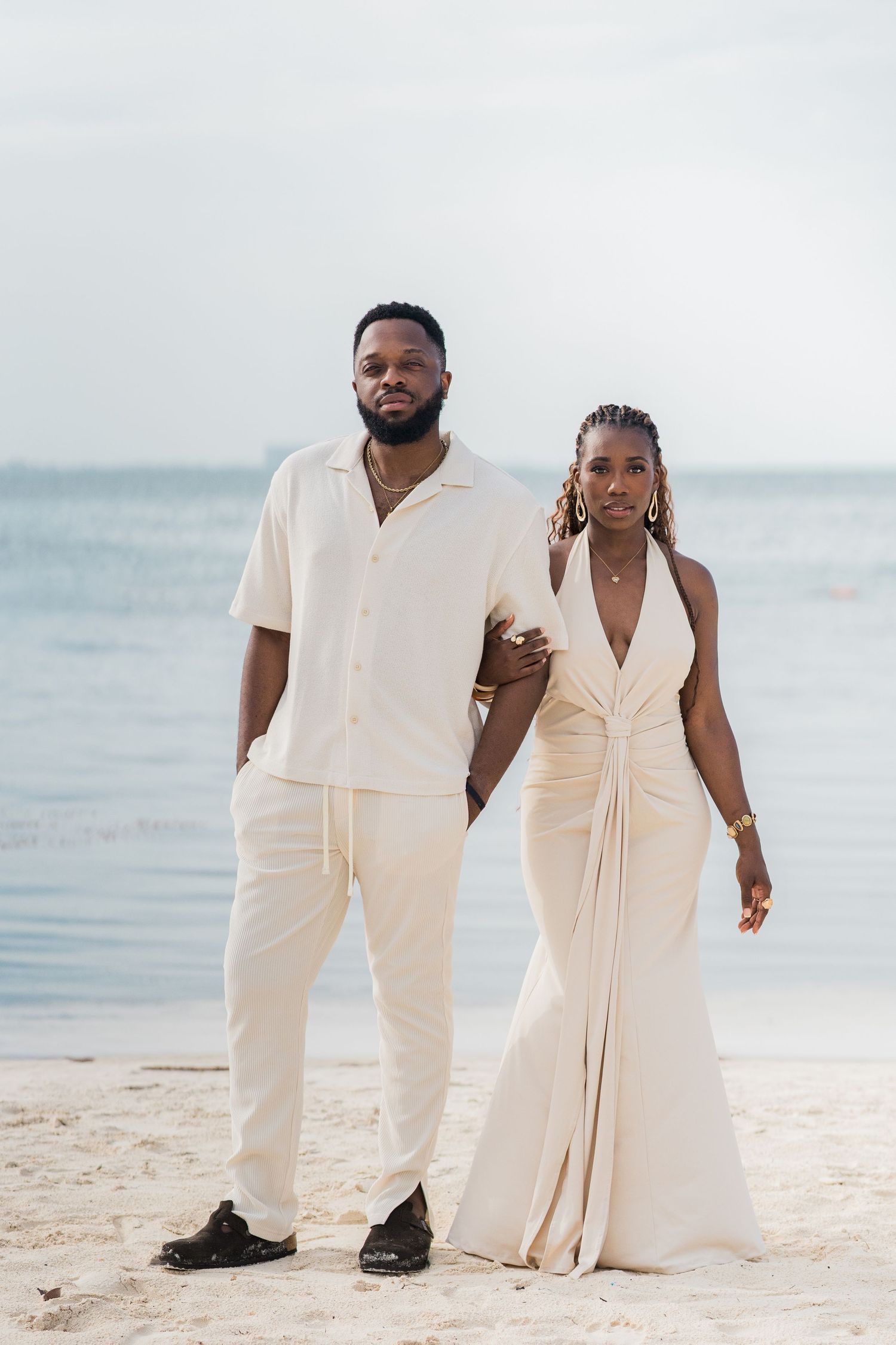 Elegant couple in matching cream outfits posing on a sandy beach with calm ocean waters in the background.