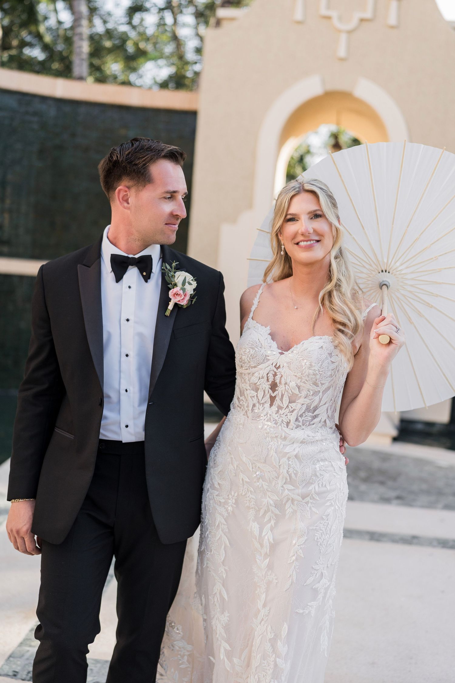 Bride in lace gown holding white parasol stands with groom in black tuxedo at outdoor wedding venue.