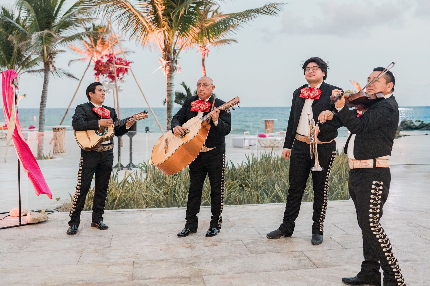 Mariachi band performing outdoors at a beachside venue, wearing traditional black and gold charro suits with red bow ties.