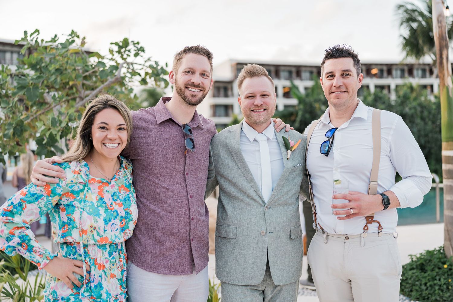 Four smiling friends pose together outdoors at a tropical wedding venue on a sunny day.