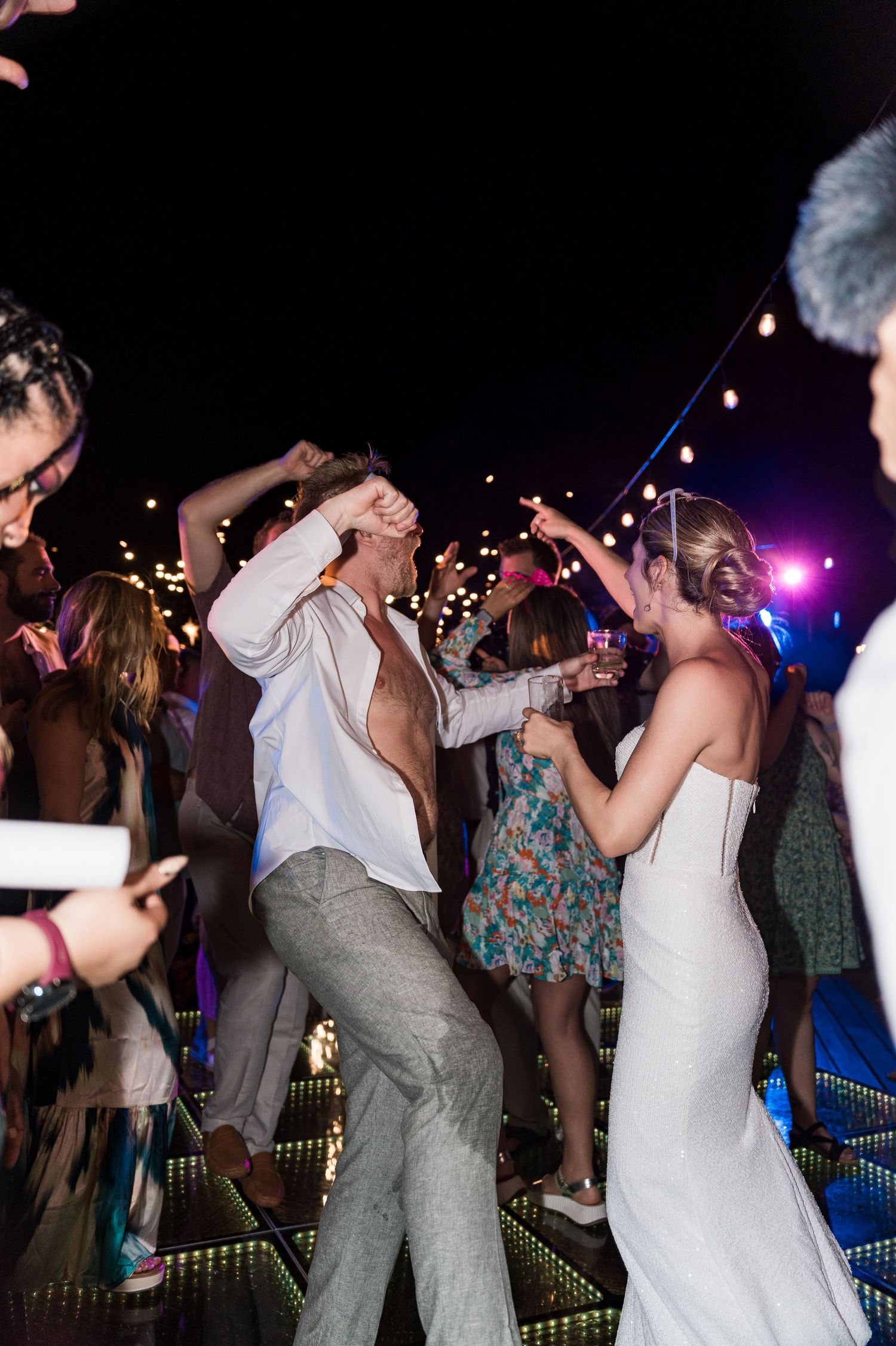 Bride and groom dancing energetically at outdoor wedding reception at night with string lights and guests surrounding them.