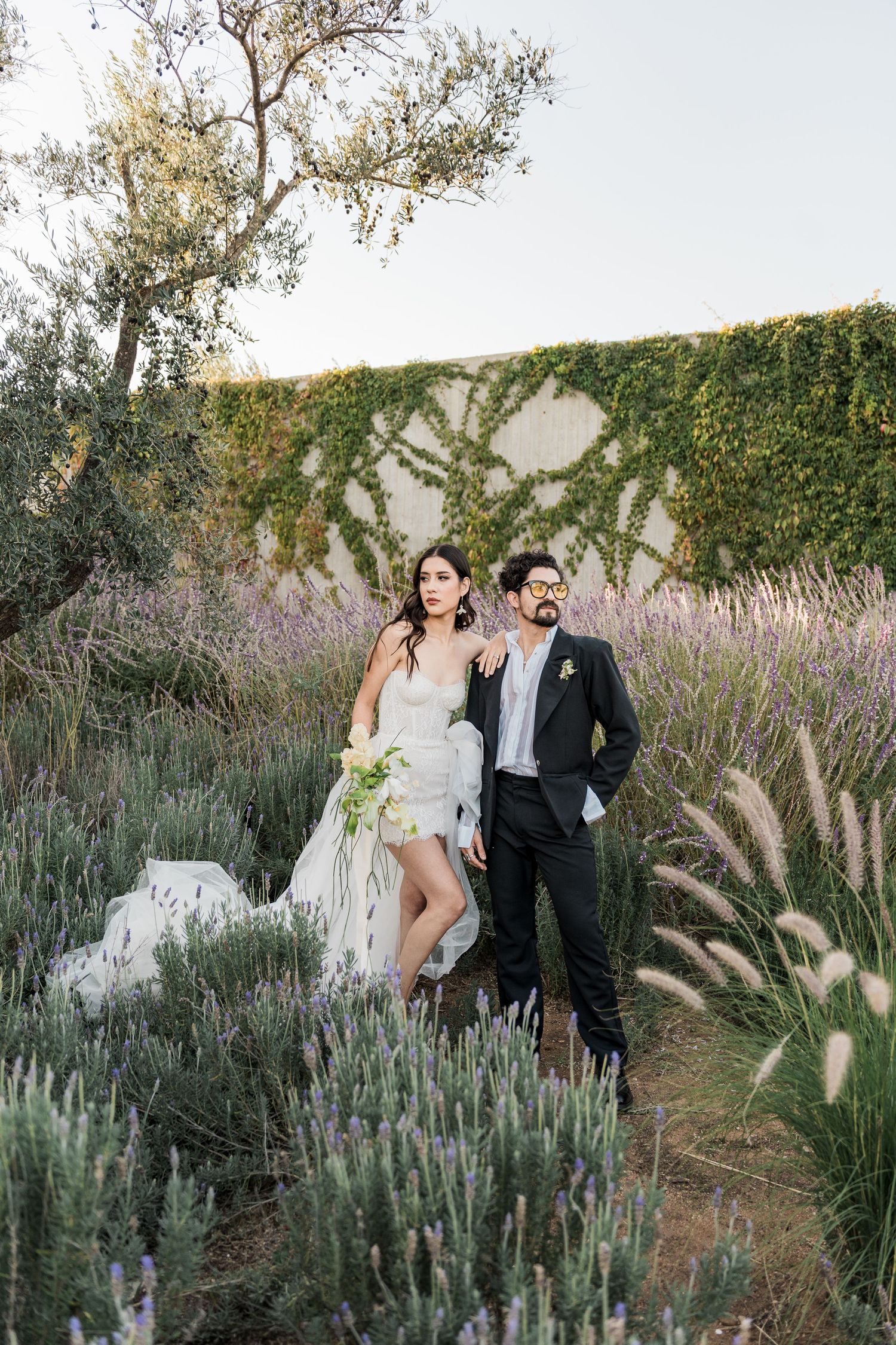 Bride and groom standing in lavender garden with ivy wall backdrop, bride holding white bouquet in elegant wedding attire. Puerto Vallarta