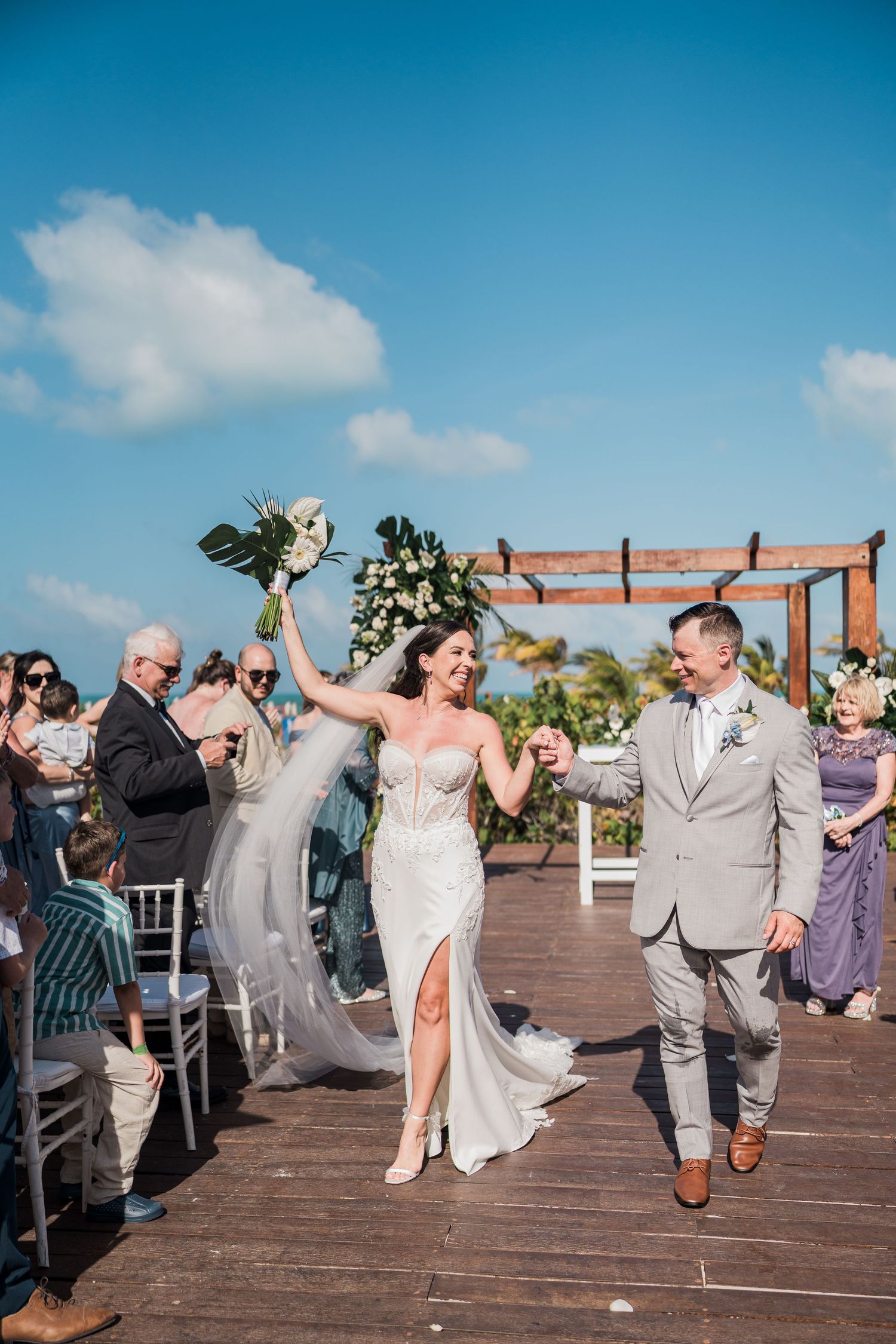 Bride and groom walk down aisle at outdoor beach wedding ceremony, bride raising bouquet in celebration under blue sky.