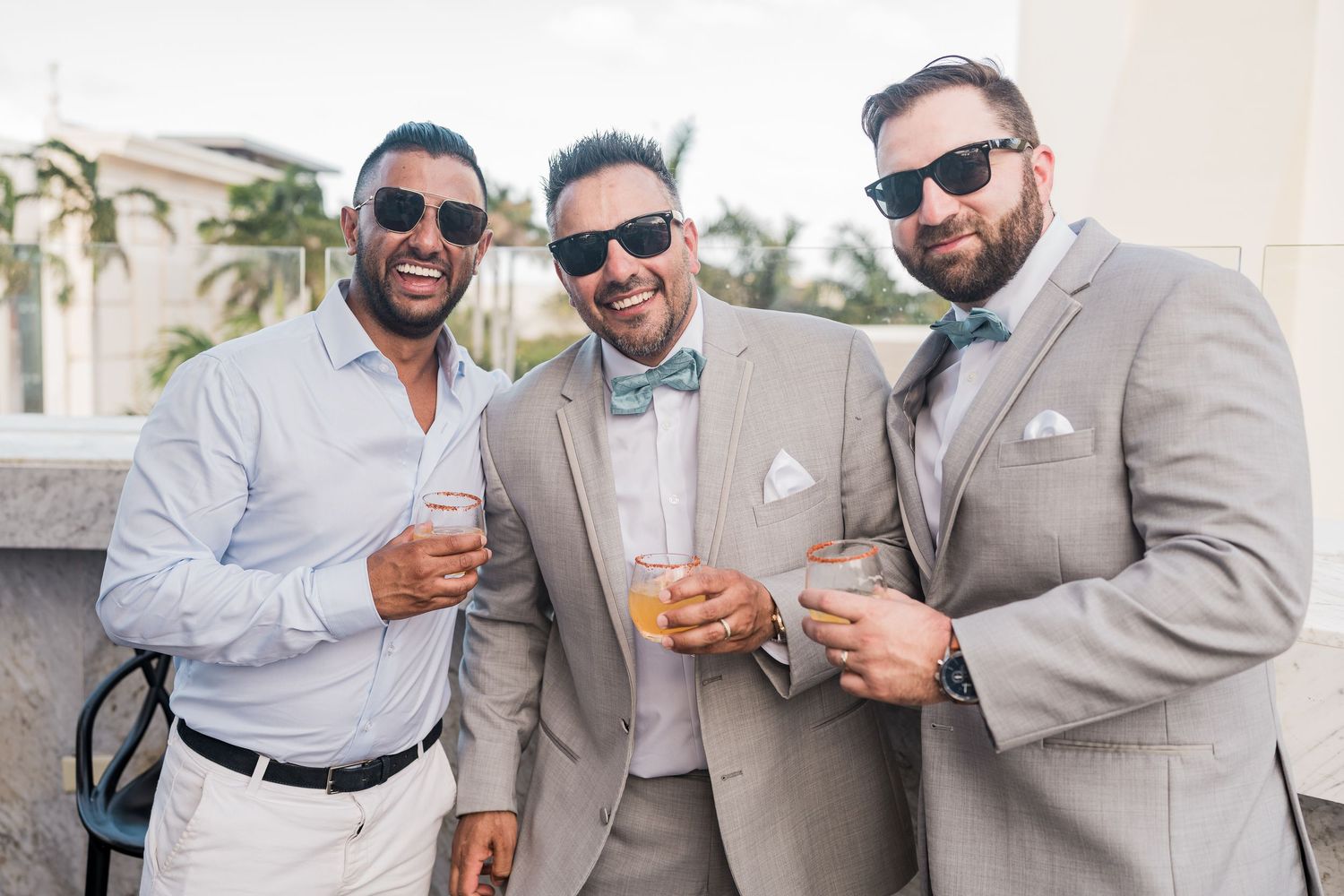 Three men in gray suits and bow ties smile while holding drinks outdoors at a formal event.