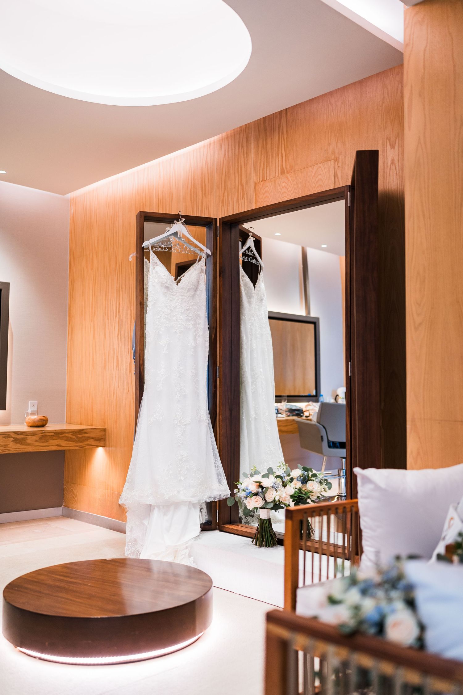 Elegant wedding dress hanging in luxury bridal suite with bouquet on wooden table.