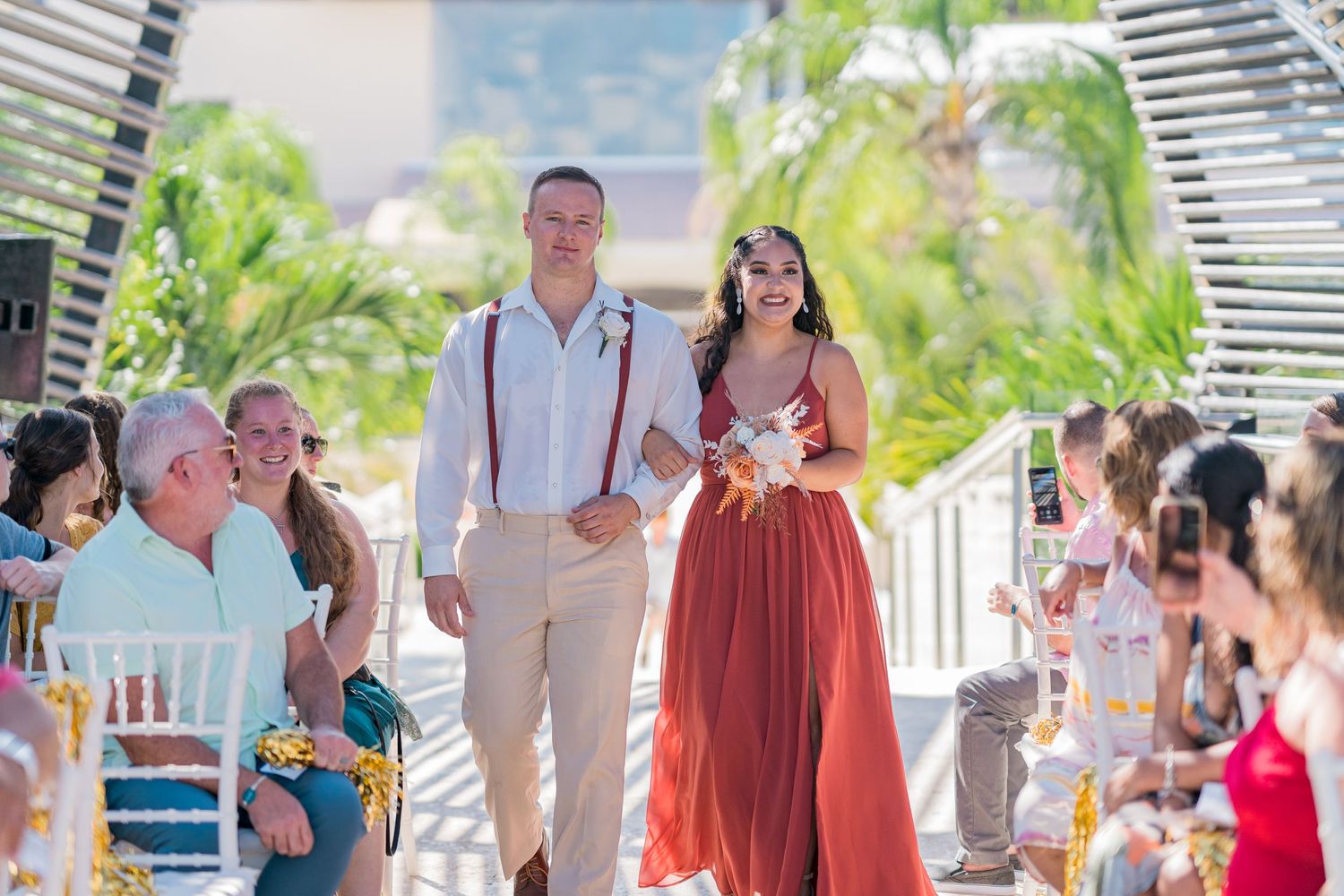 Groomsman in suspenders walks bridesmaid in red floral dress down outdoor wedding ceremony aisle.