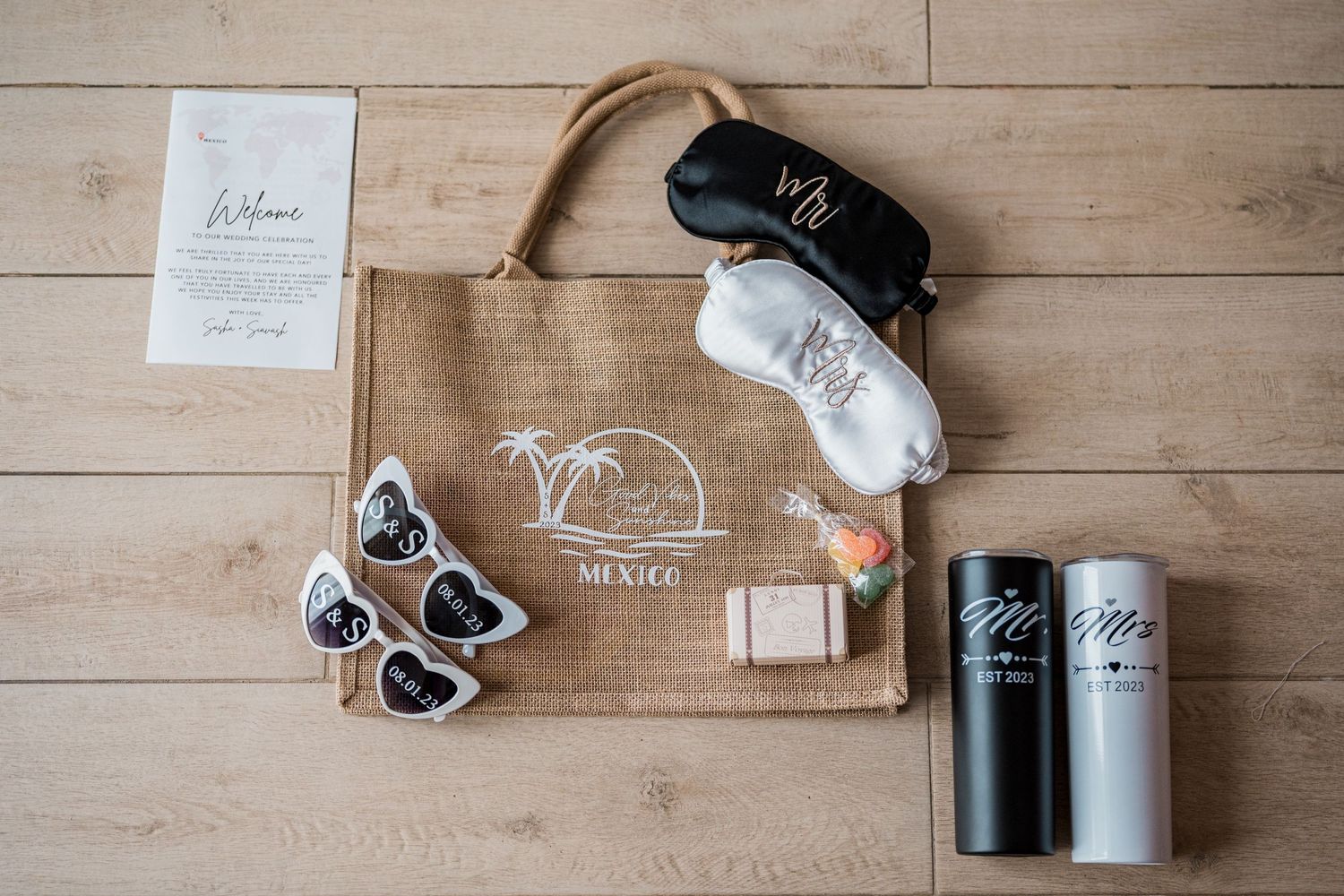 Wedding welcome gift bag with sleep masks, tumblers, sunglasses, and soap on a wooden floor.