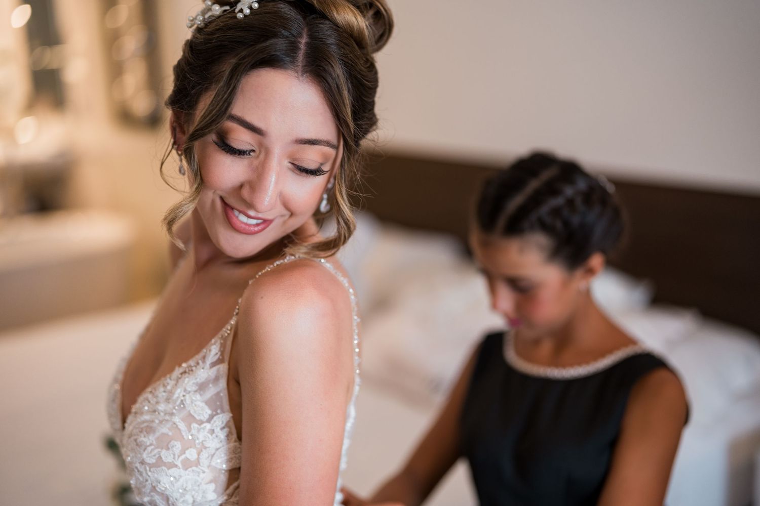 Bride smiling as bridesmaid fastens back of lace wedding dress.