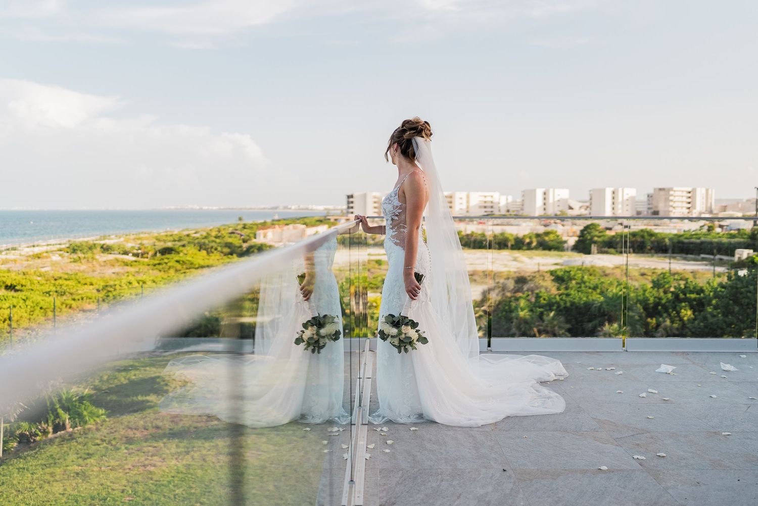 Bride in white gown with flowing veil stands on rooftop terrace overlooking coastal cityscape.