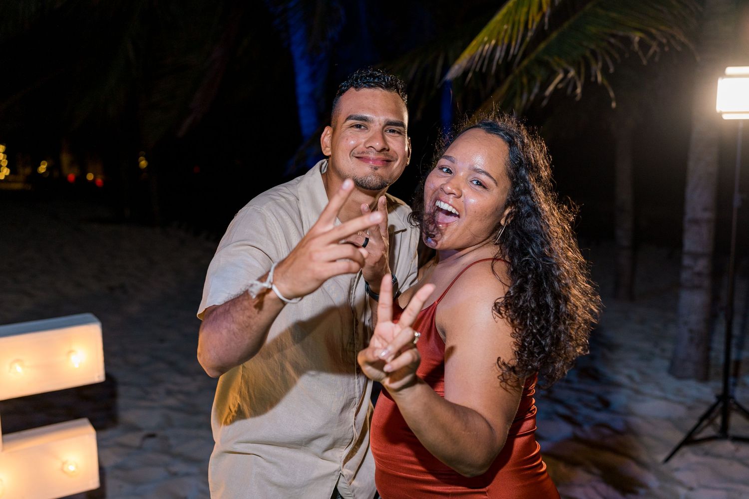 Smiling couple making peace signs together at tropical beach wedding reception at night.