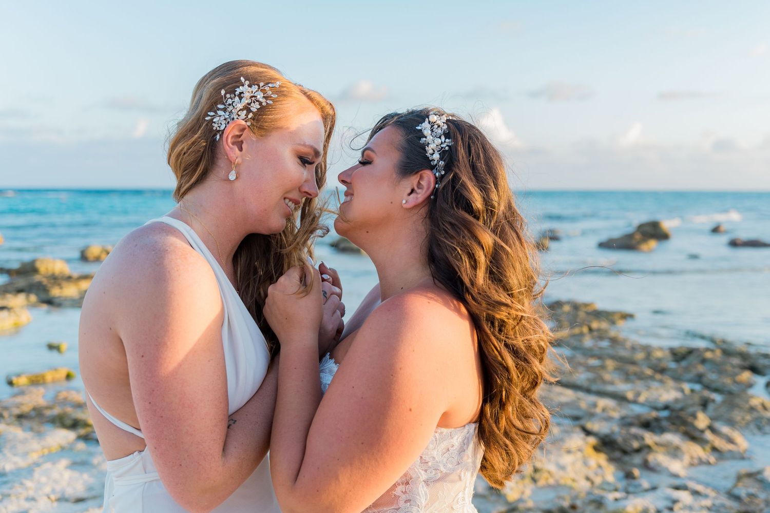 Two brides touching foreheads tenderly by a rocky ocean shore at a beach wedding.