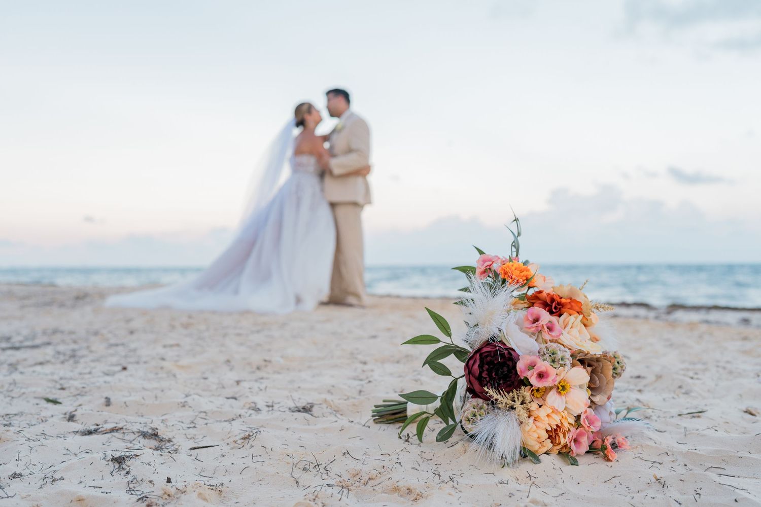 Bridal bouquet with coral and burgundy flowers in foreground with couple embracing on beach.