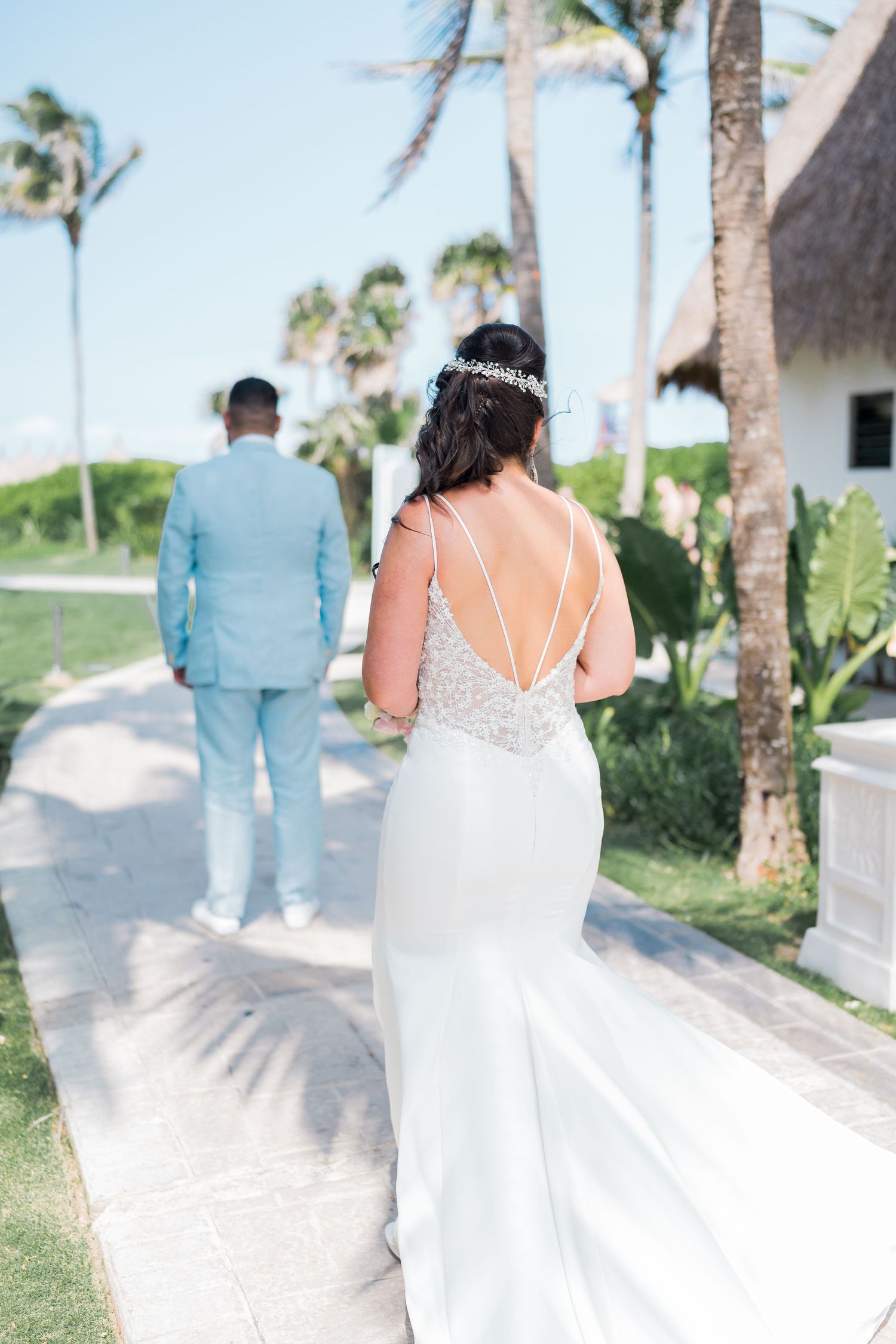 Bride in a backless white gown approaching groom in light blue suit for a first look outdoors.
