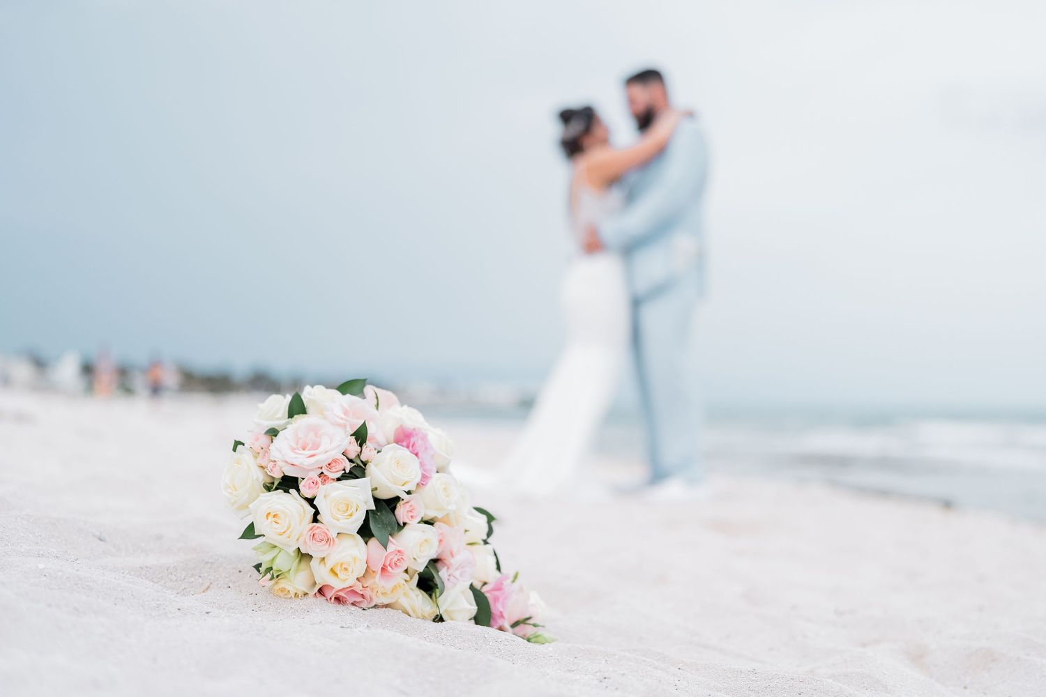 Bridal bouquet of pink and white roses on white sand with couple embracing in the background.