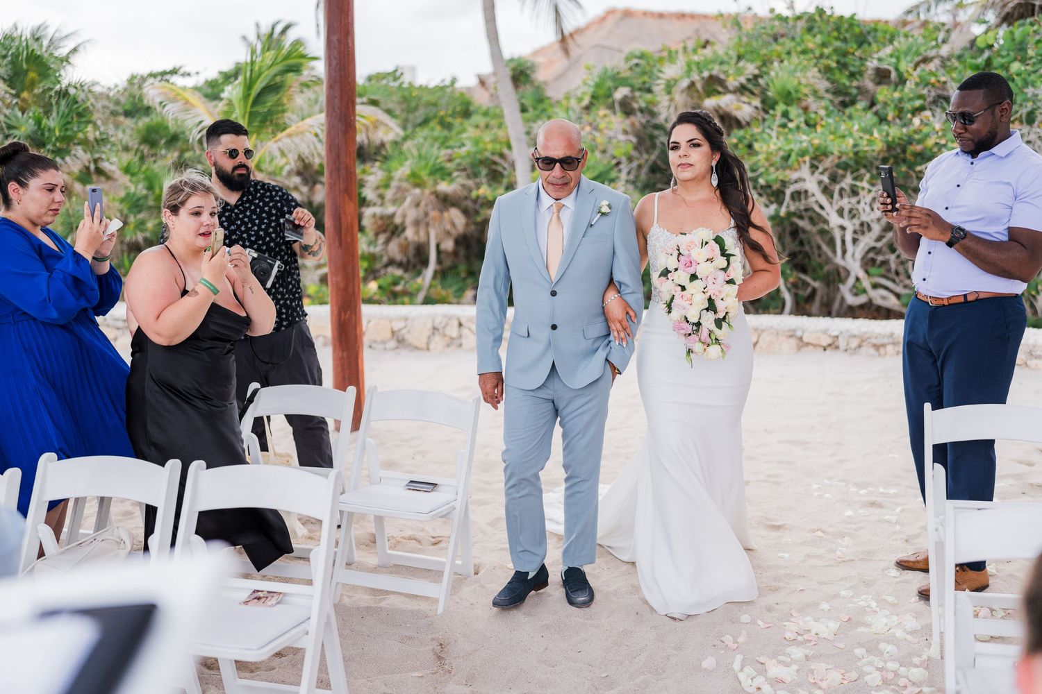 Father walks bride down sandy aisle as guests applaud at tropical beach wedding ceremony.
