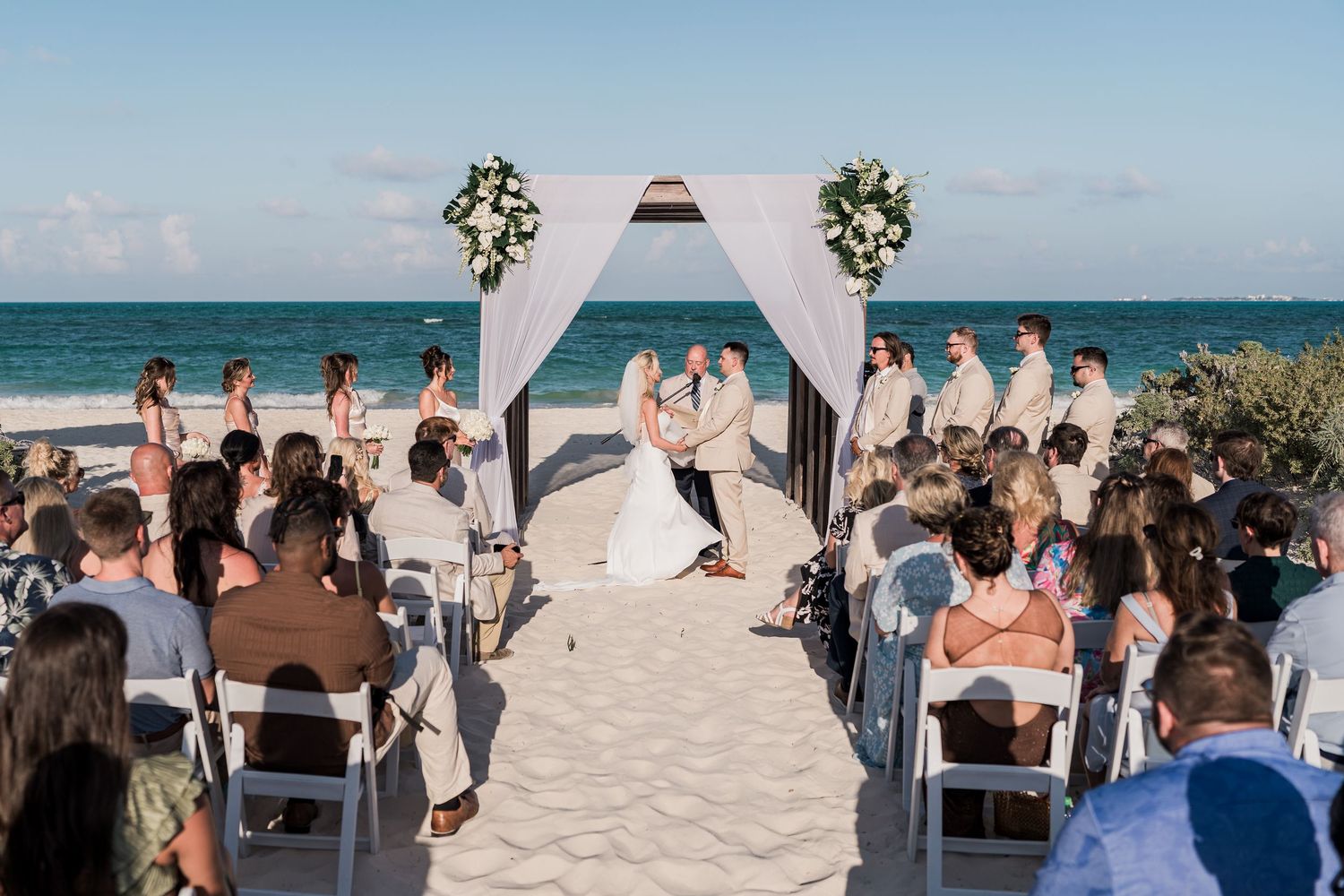 Beach wedding ceremony with couple at floral arch altar, ocean backdrop, guests seated on white chairs in sand.
