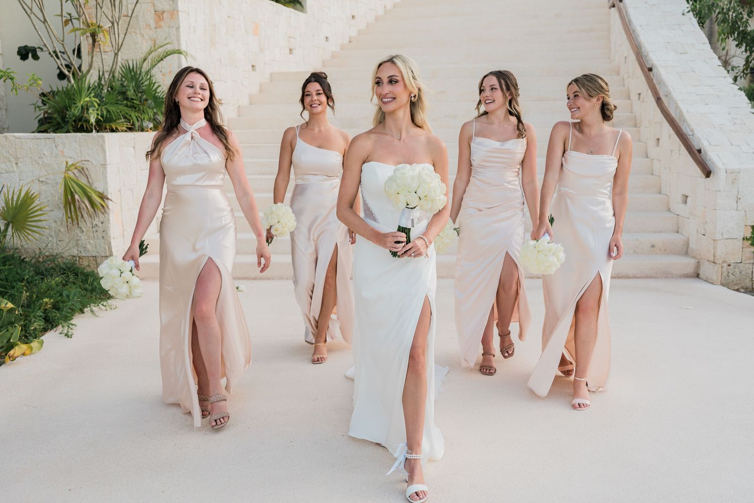 Bride in white dress walks confidently with four bridesmaids in blush gowns at a tropical wedding venue.