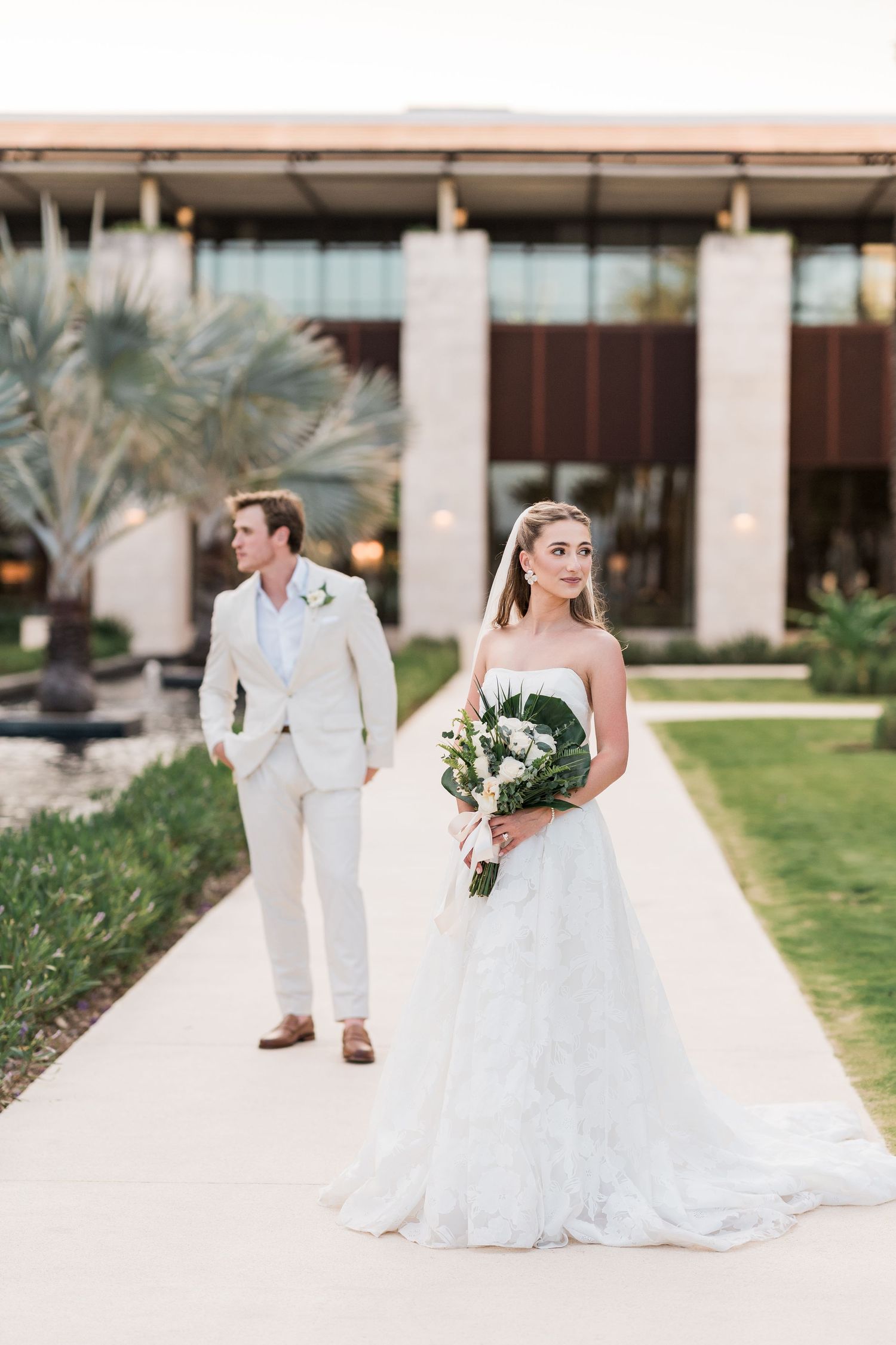 Bride holding bouquet in lace gown with groom in tan suit outdoors.