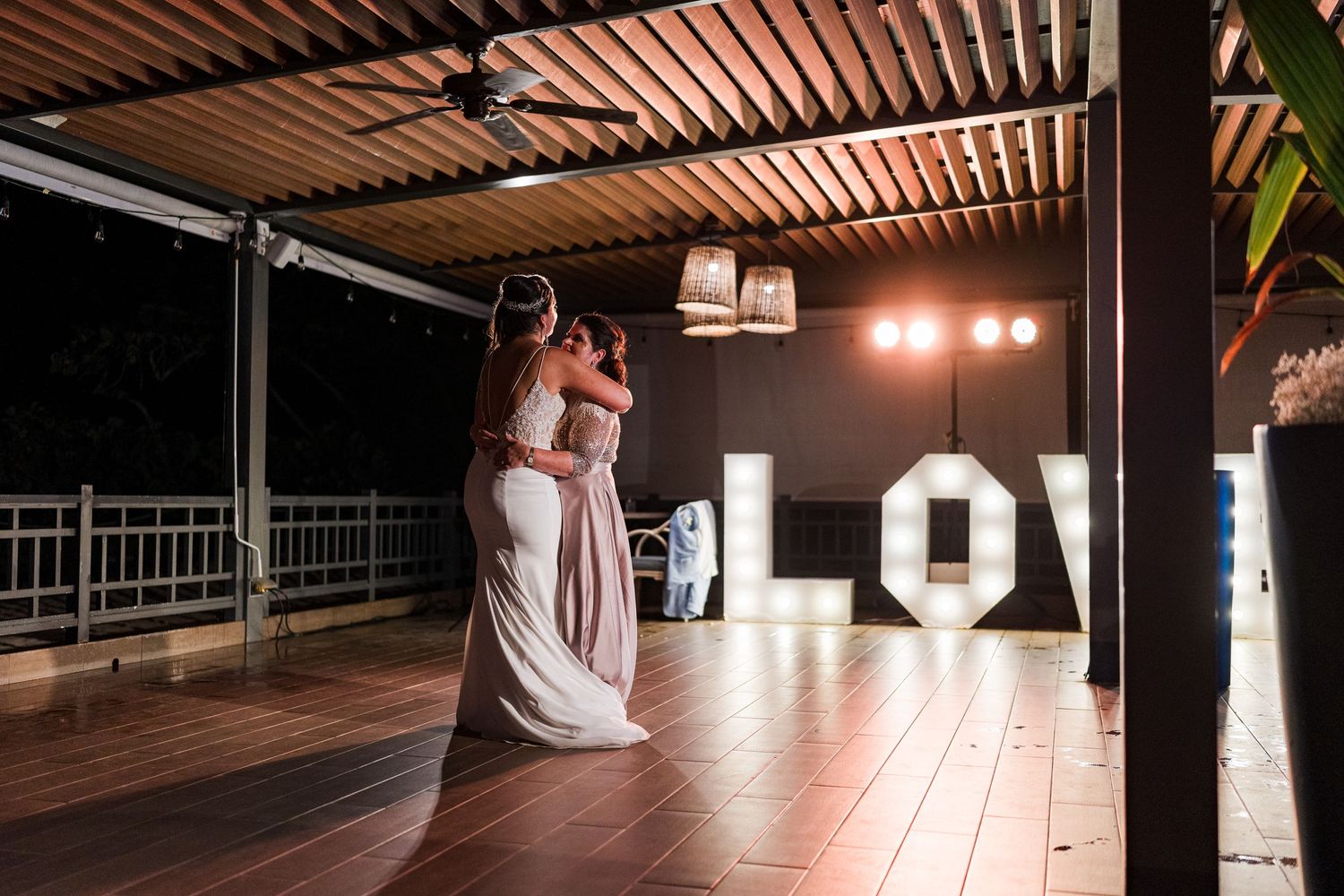 Bride sharing a tender embrace with a bridesmaid near illuminated LOVE letters at night wedding.