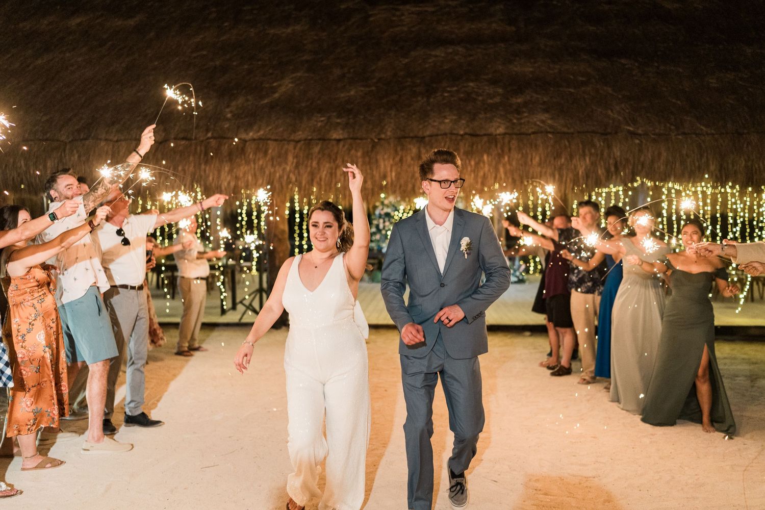Newlyweds walking through sparkler exit tunnel at romantic outdoor beach wedding reception.