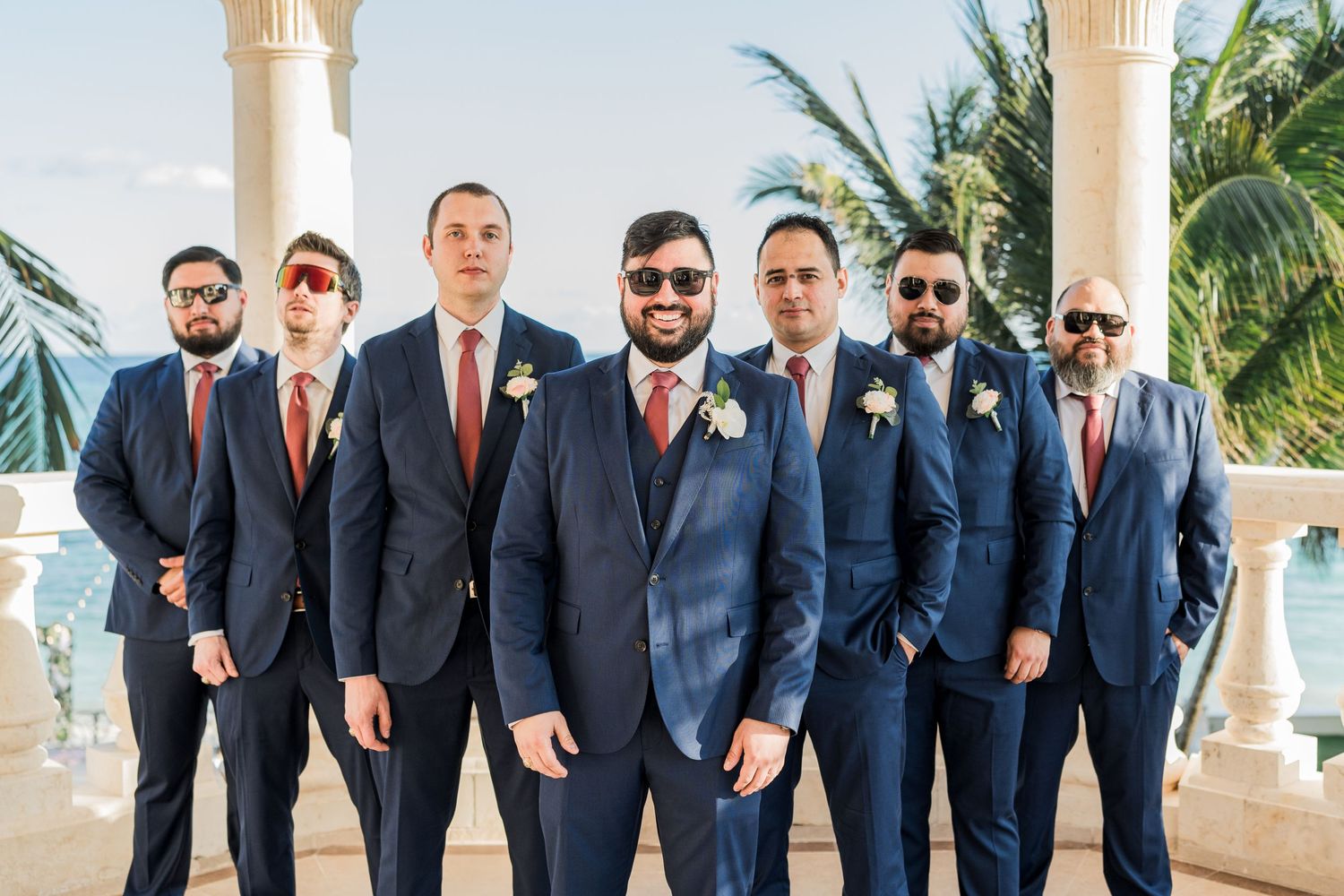 Groom and six groomsmen in navy suits with burgundy ties posing at sunlit oceanfront wedding venue with columns.