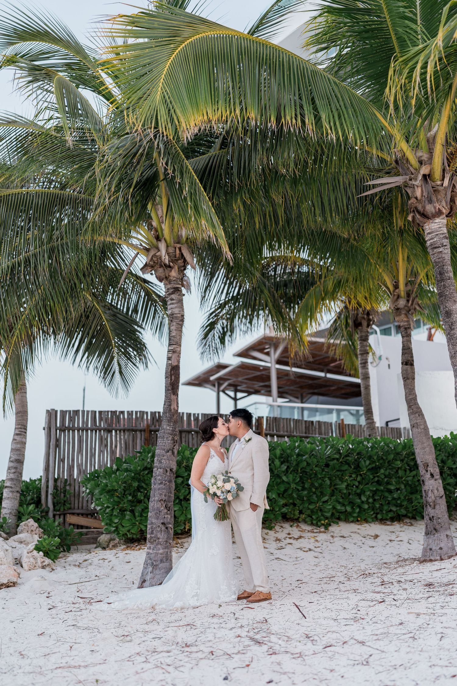 Newlyweds share a kiss beneath tropical palm trees on a white sandy beach at their destination wedding.