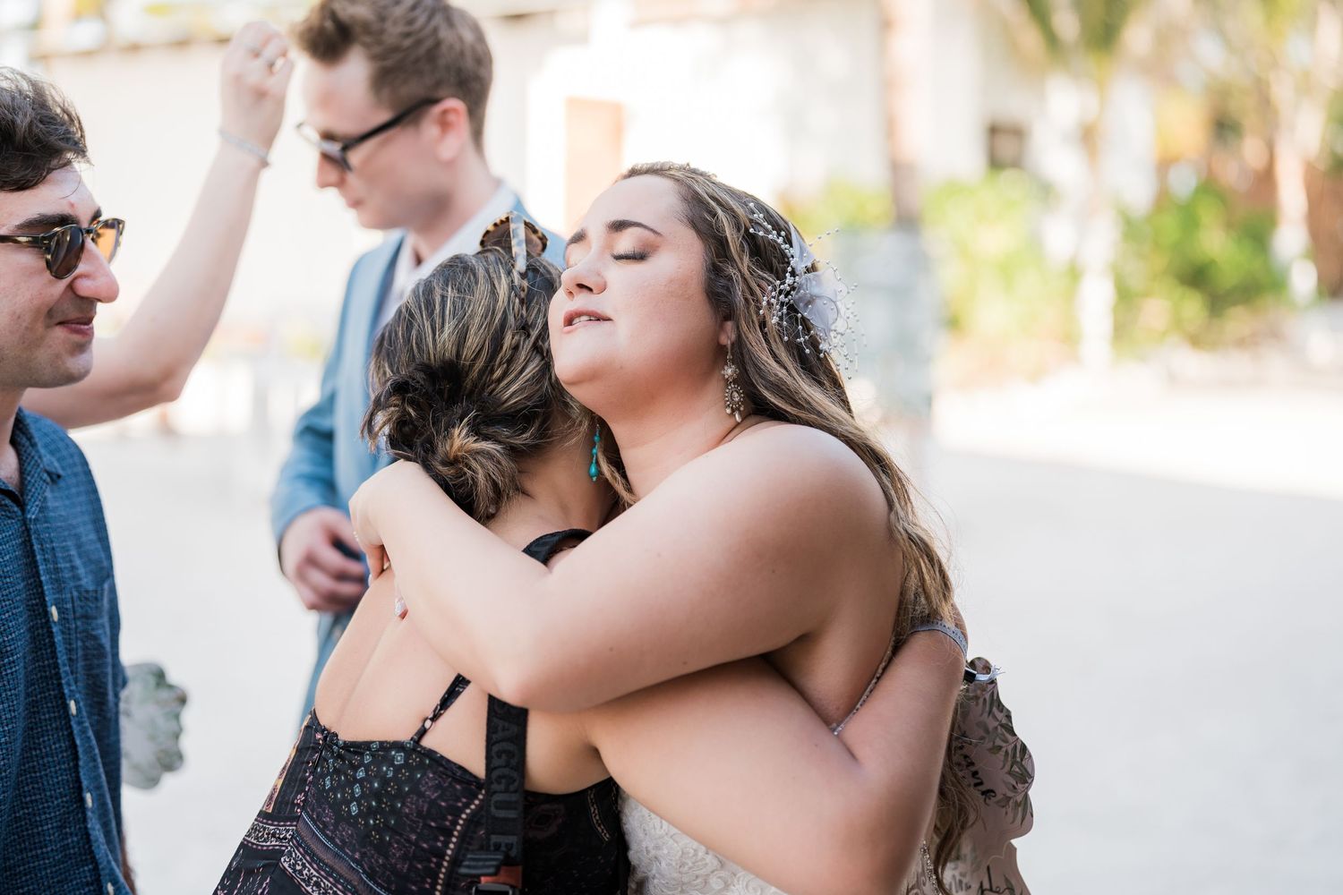Bride in lace wedding dress shares emotional hug with guest at tropical beach wedding.