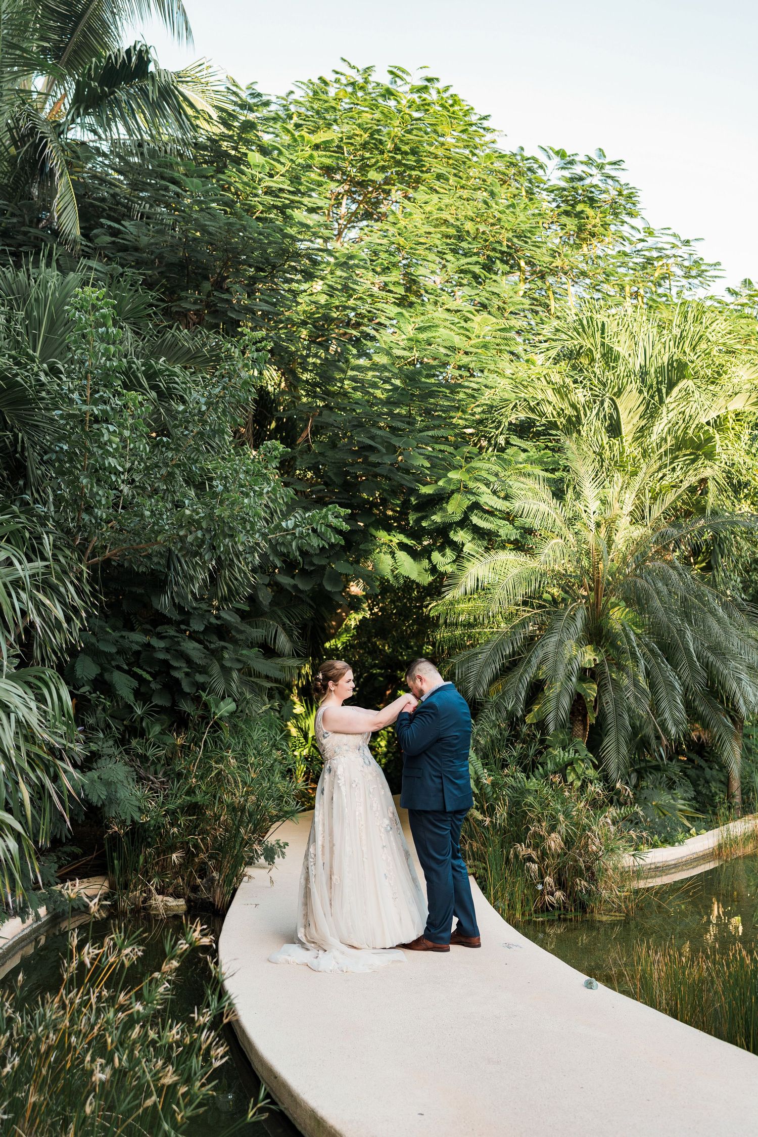 Secrets Akumal Wedding - Bride and groom share a romantic moment on a curved bridge surrounded by lush tropical greenery and a serene pond.