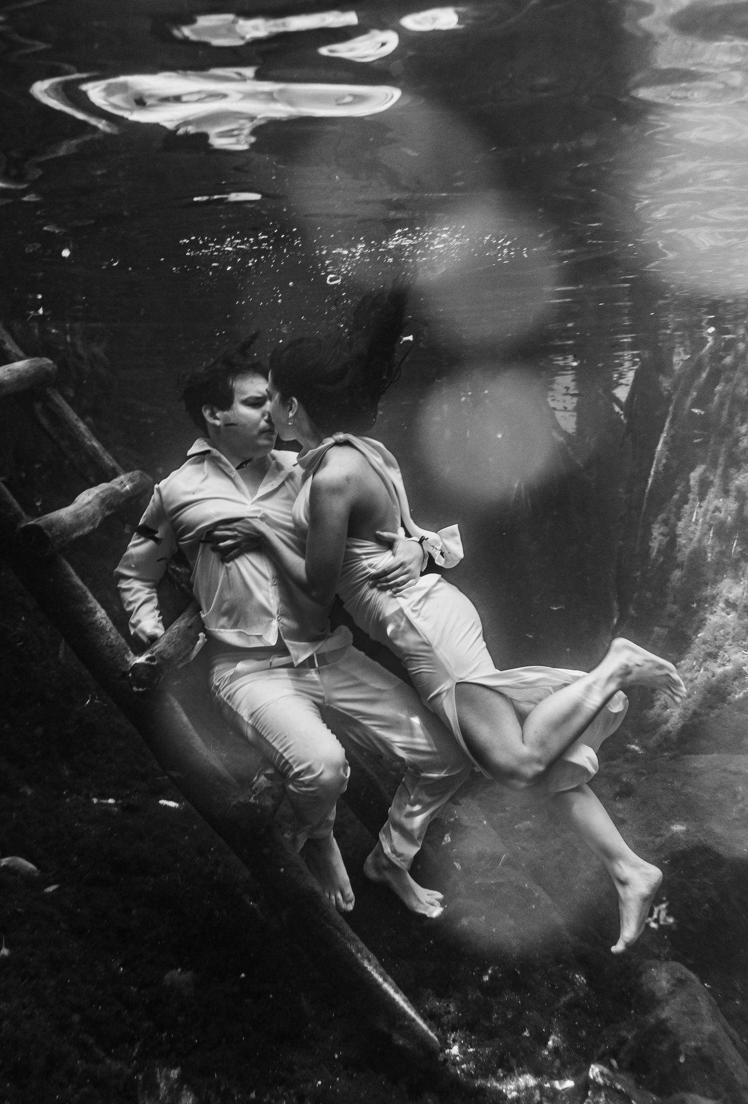 Black and white underwater photo of a couple embracing romantically near wooden logs in clear water.
