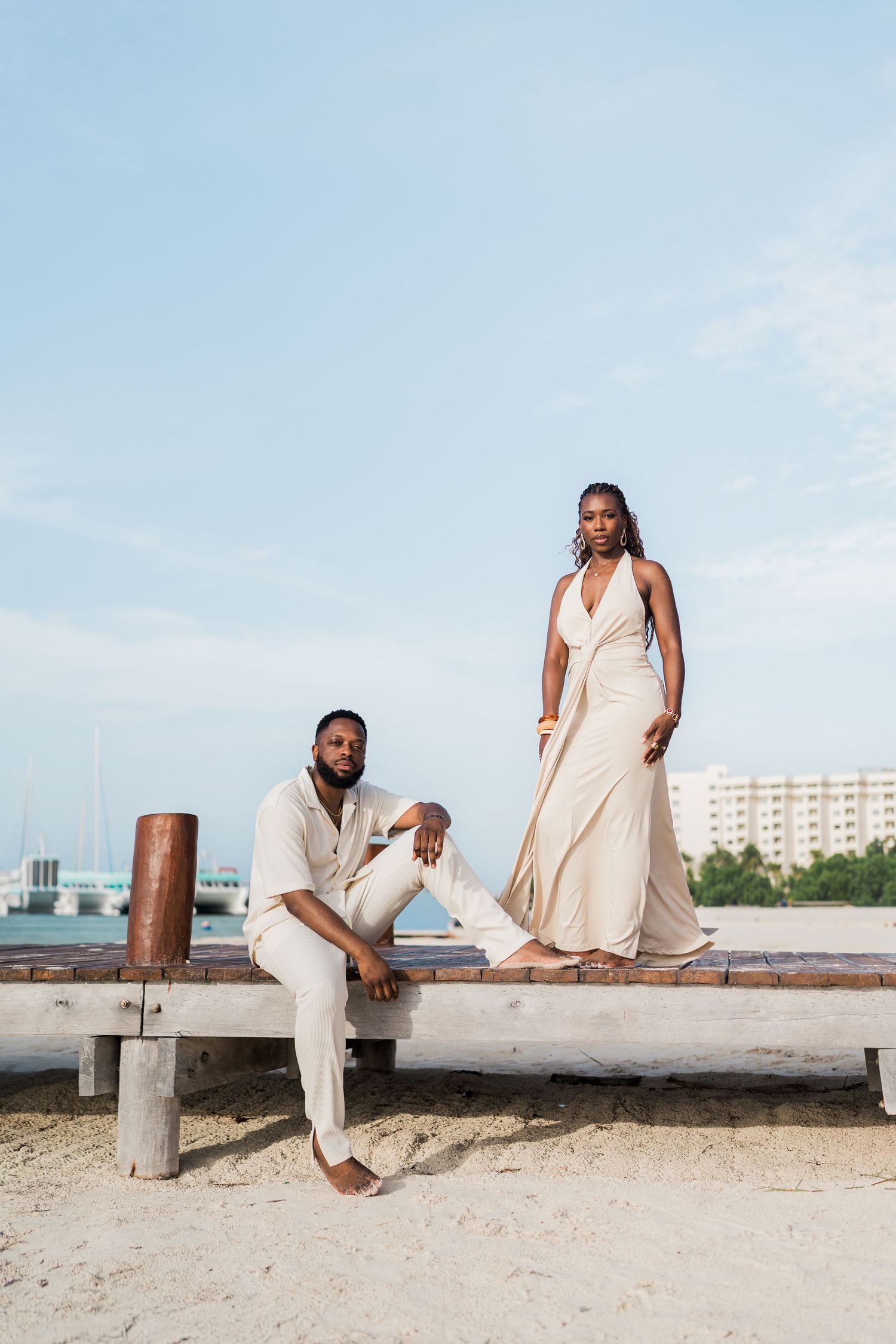 A Black couple in matching cream outfits pose on a wooden beach dock, man seated and woman standing elegantly.